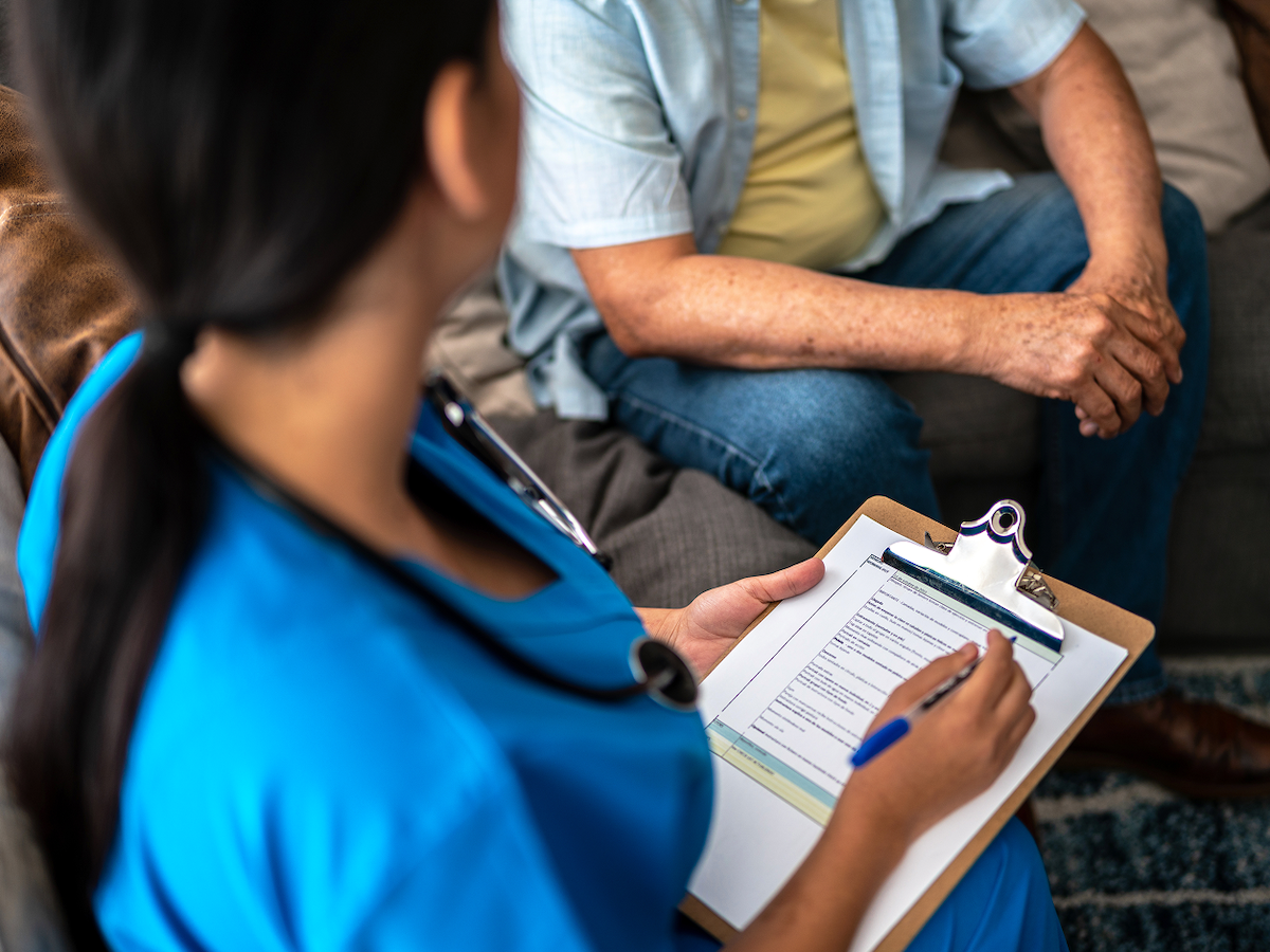 Photo of a nurse holding a clipboard and talking to a member on a sofa