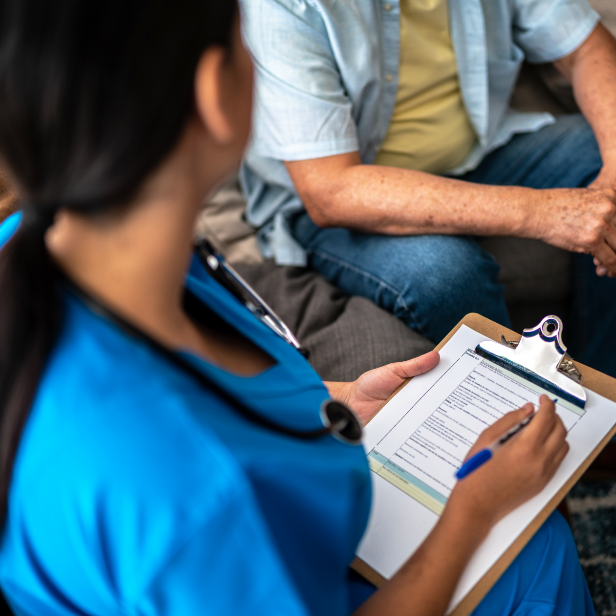 Photo of a nurse holding a clipboard and pen while speaking to a member on a sofa