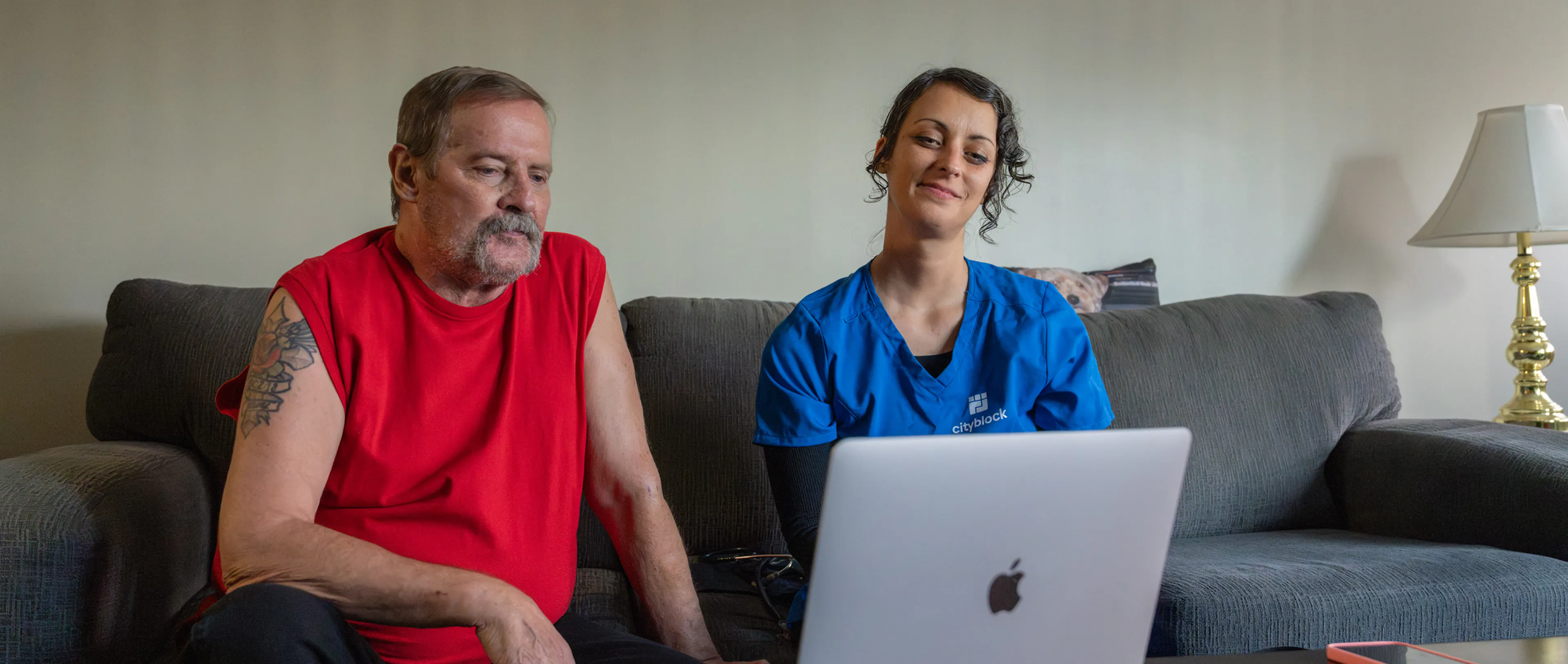 Photo of an older member and a Cityblock care team member in blue scrubs sitting on a couch, both looking at a laptop.