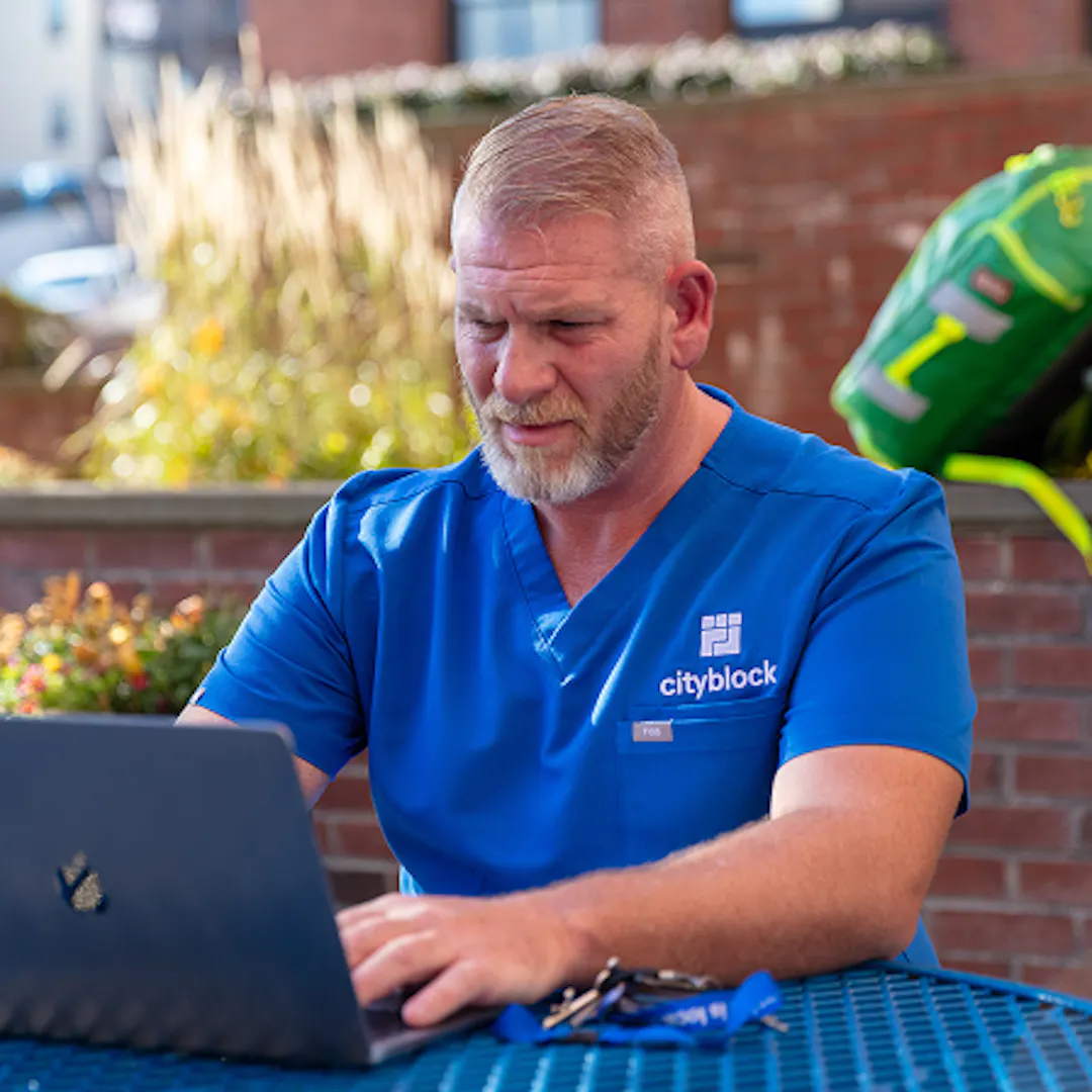 Photo of a care team member with a beard wearing blue Cityblock scrubs, sitting at an outdoor table and focused on working on a laptop.