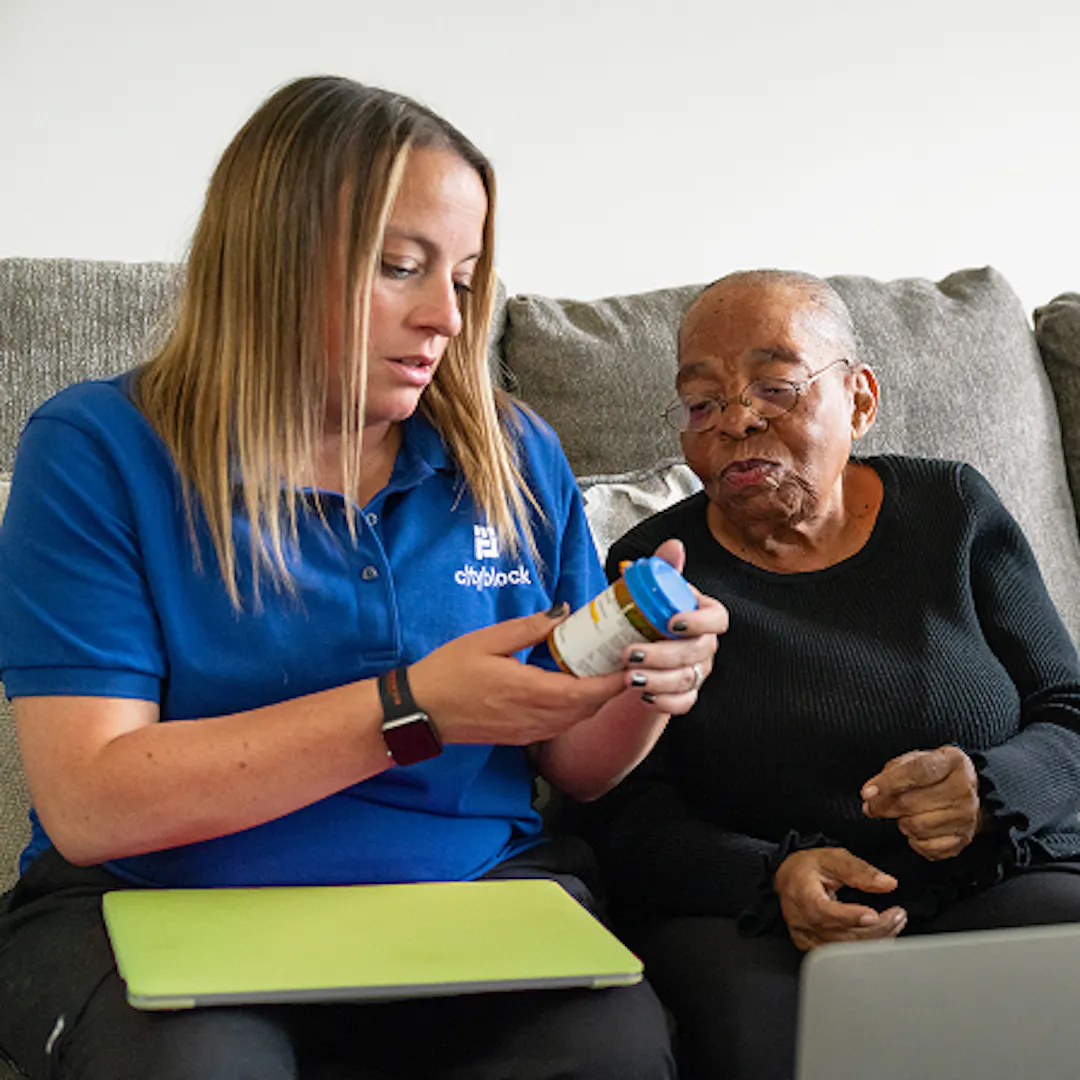 Photo of a care team member in a blue Cityblock polo shirt reviewing a prescription medication bottle with an older member on a couch.