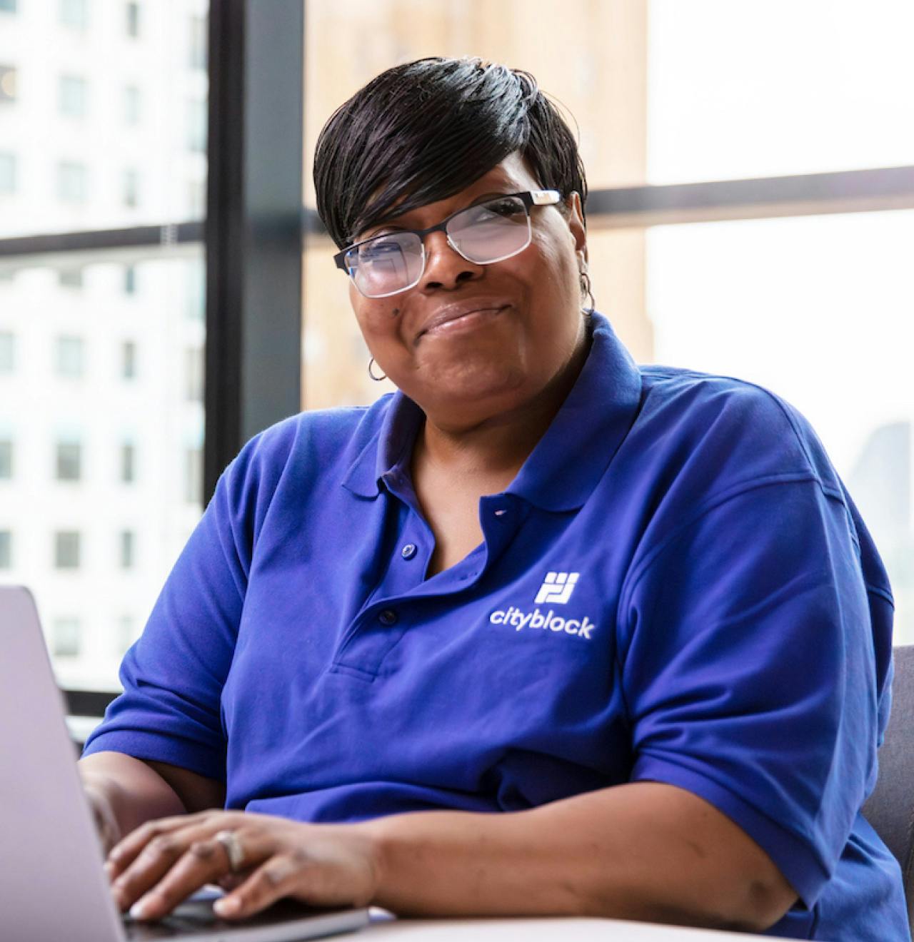 Woman wearing blue Cityblock polo and glasses smiles