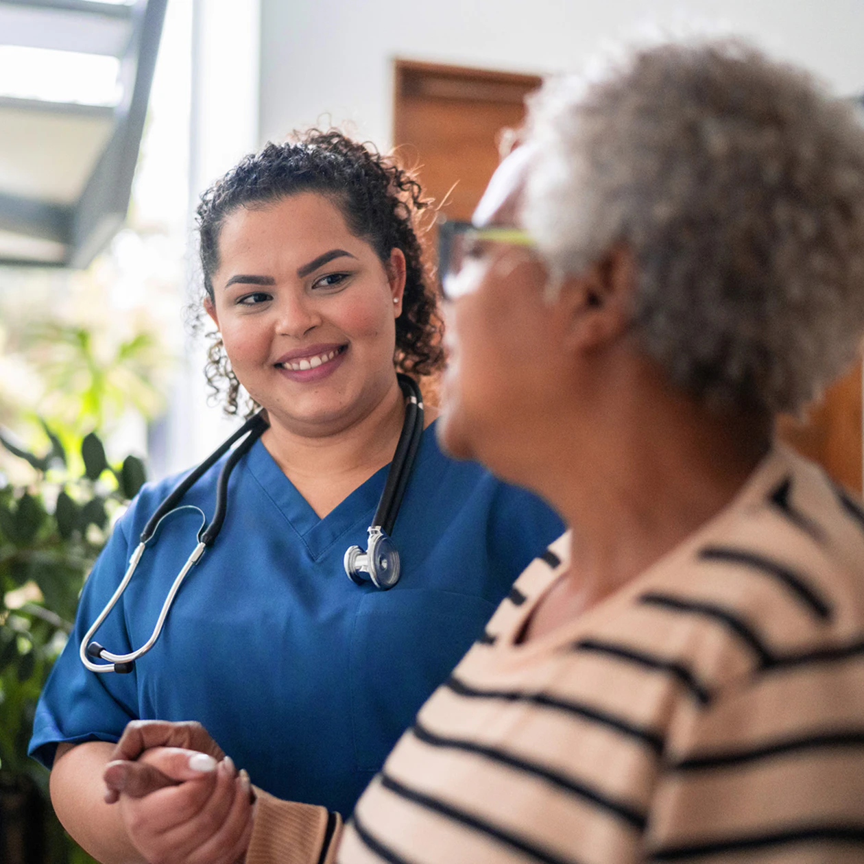 Image of a smiling nurse in blue scrubs gently holding hands with an older female member during a medical visit.