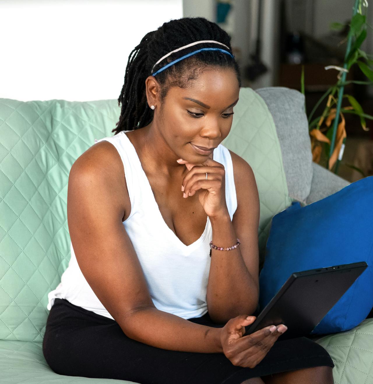 Woman wearing white tank sitting on the couch smiling while looking at a computer tablet.