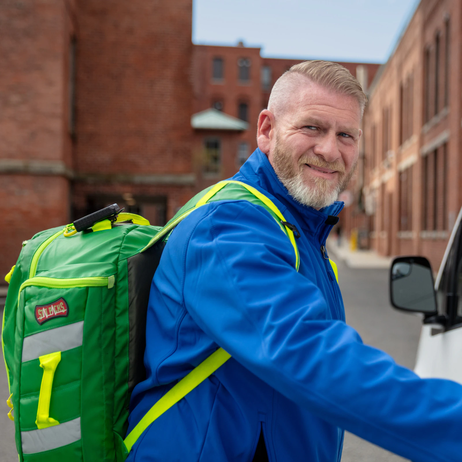Image of a smiling clinician wearing a blue jacket and carrying a bright green backpack outdoors near a vehicle.