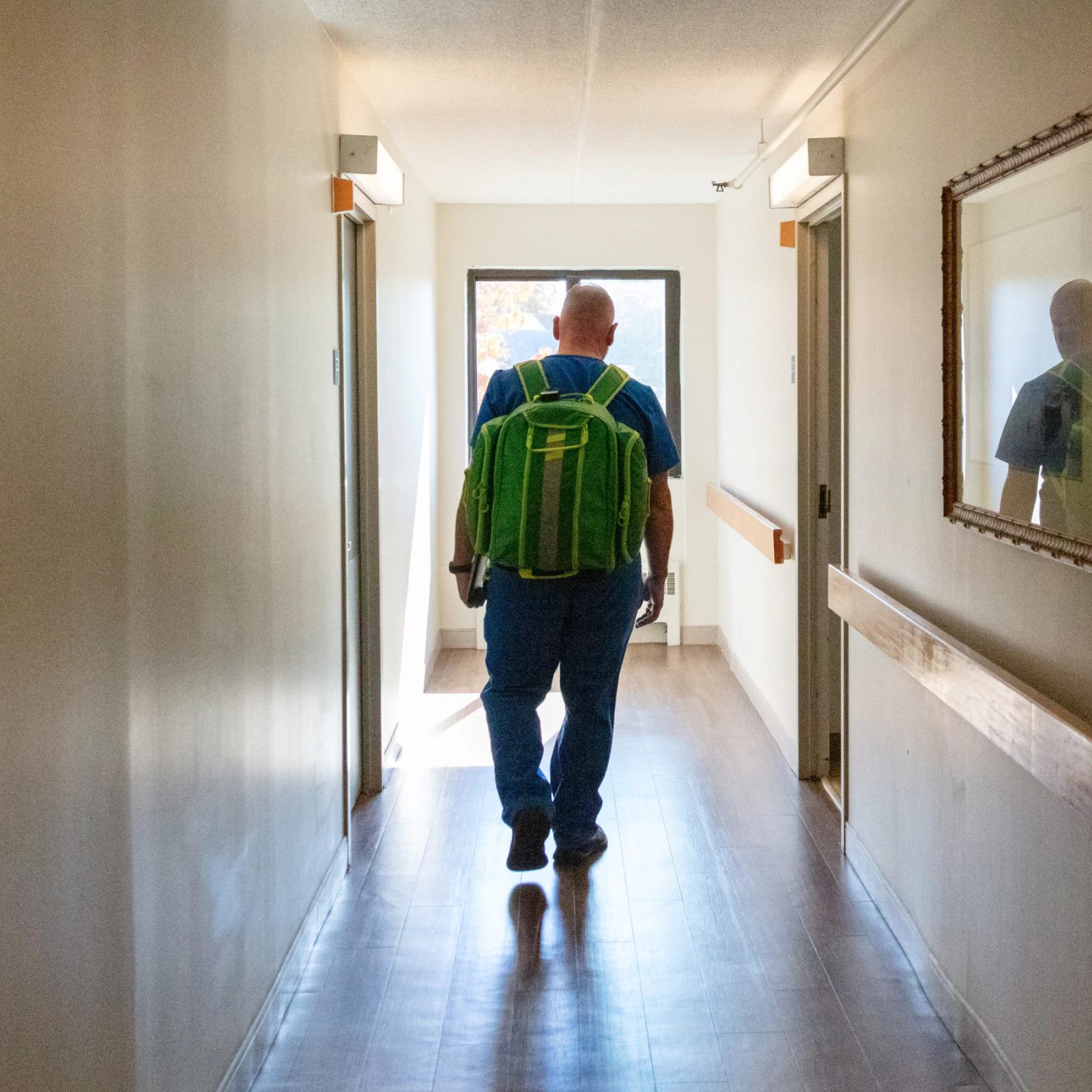 Image of a clinician wearing blue scrubs and a large green backpack walking down a bright hallway toward a window.