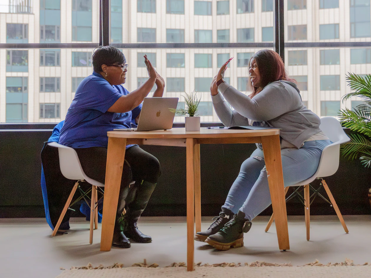Image of a community health partner and member high-fiving across a round wooden table with a laptop, sitting indoors near a large window.