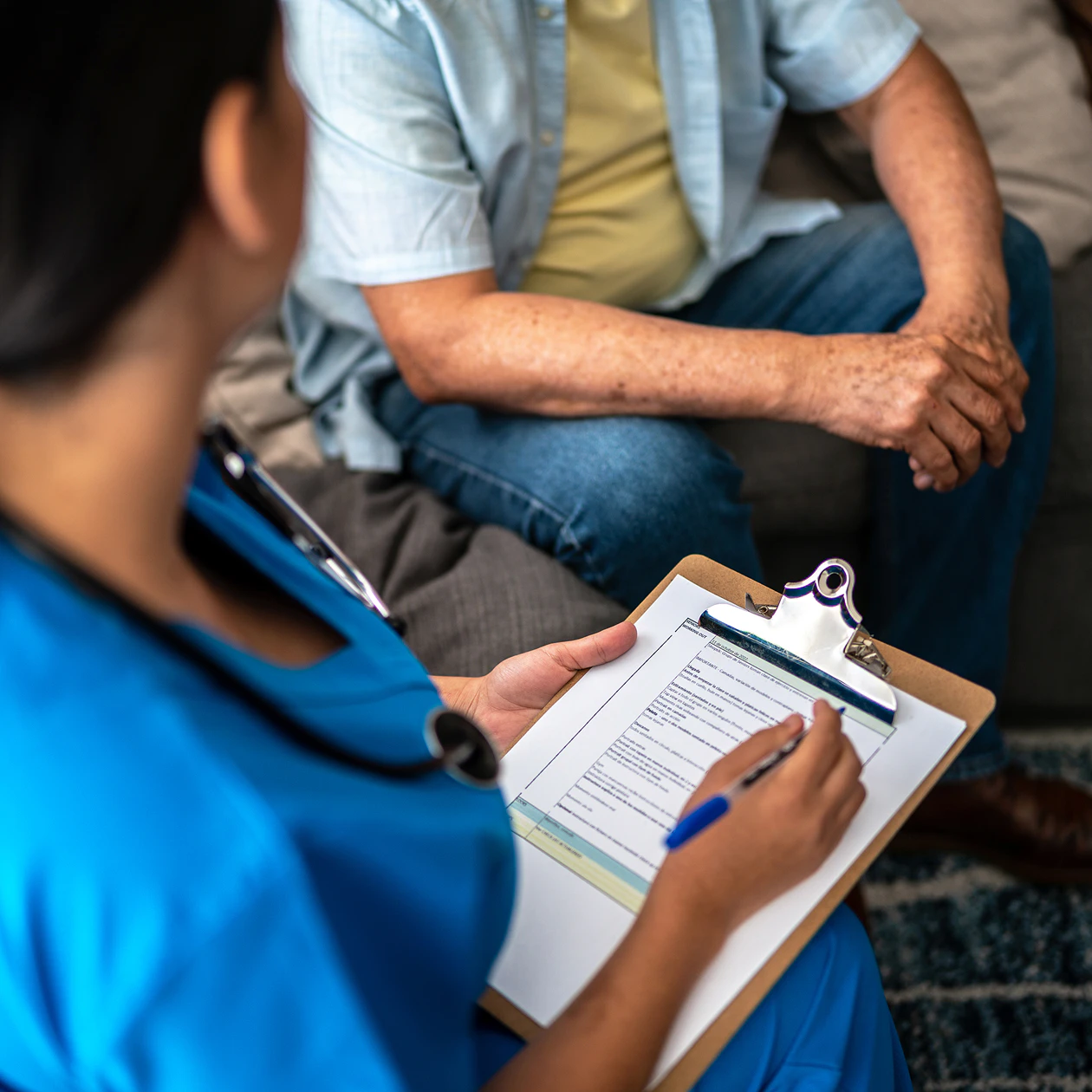 Image of a nurse in blue scrubs writing on a clipboard while consulting with an older man seated on a couch.