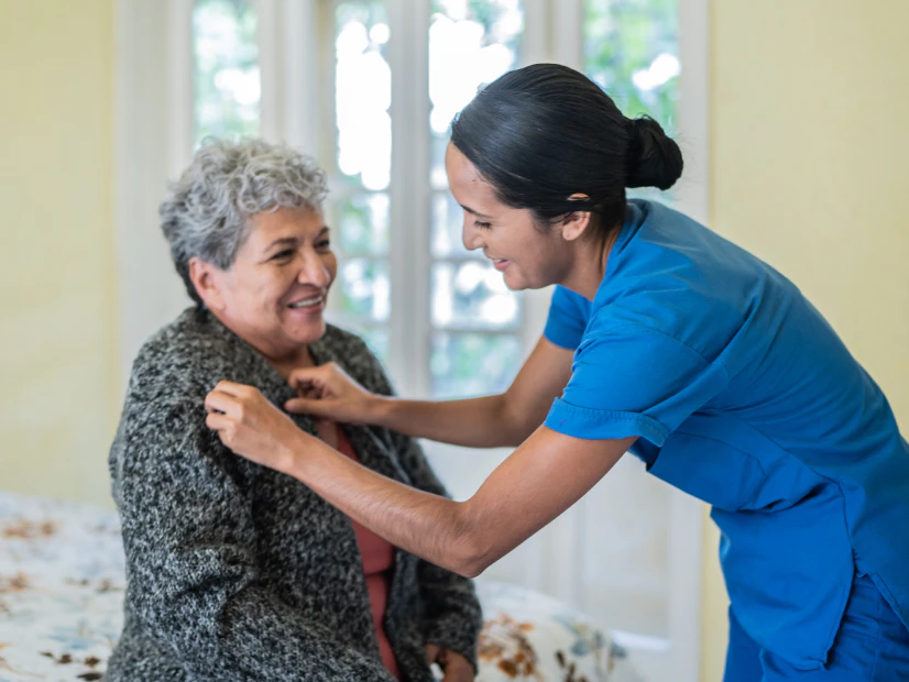 Image of a female caregiver in blue scrubs helping an older woman adjust her gray sweater in a bedroom.