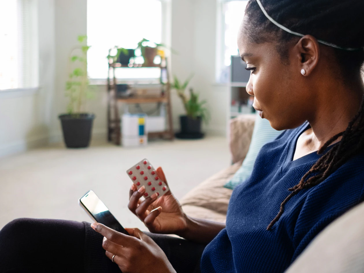 Image of a woman sitting on a couch, holding a smartphone in one hand and a blister pack of pills in the other.