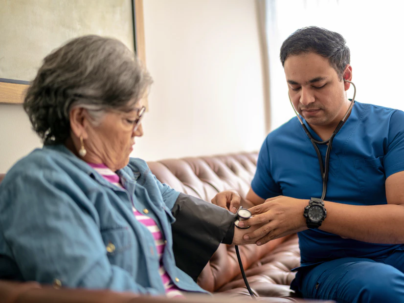 Image of a male healthcare worker in blue scrubs using a stethoscope to take an older woman's blood pressure on a couch.