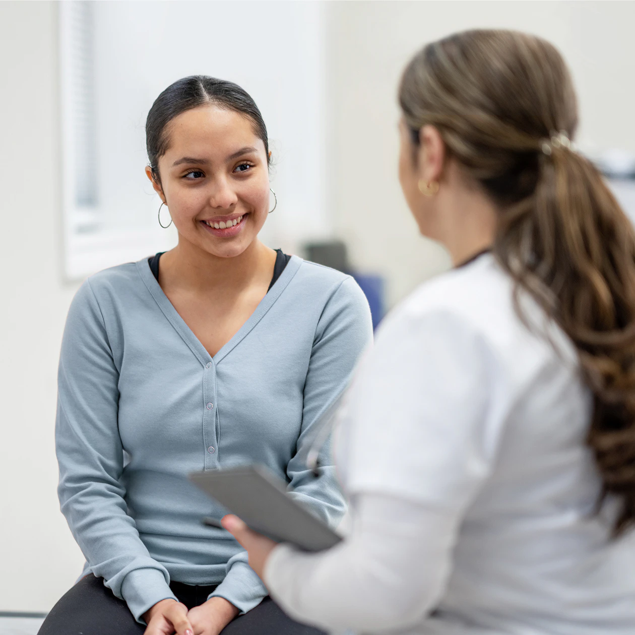 Image of a young female member smiling while sitting and speaking with a medical provider holding a tablet in a clinic.