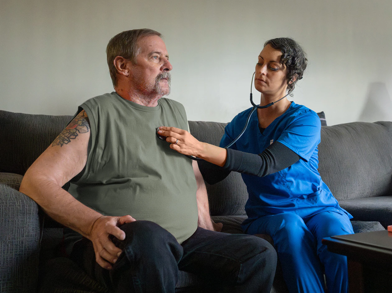 Image of a nurse in blue scrubs using a stethoscope to listen to the heart of a seated older man.
