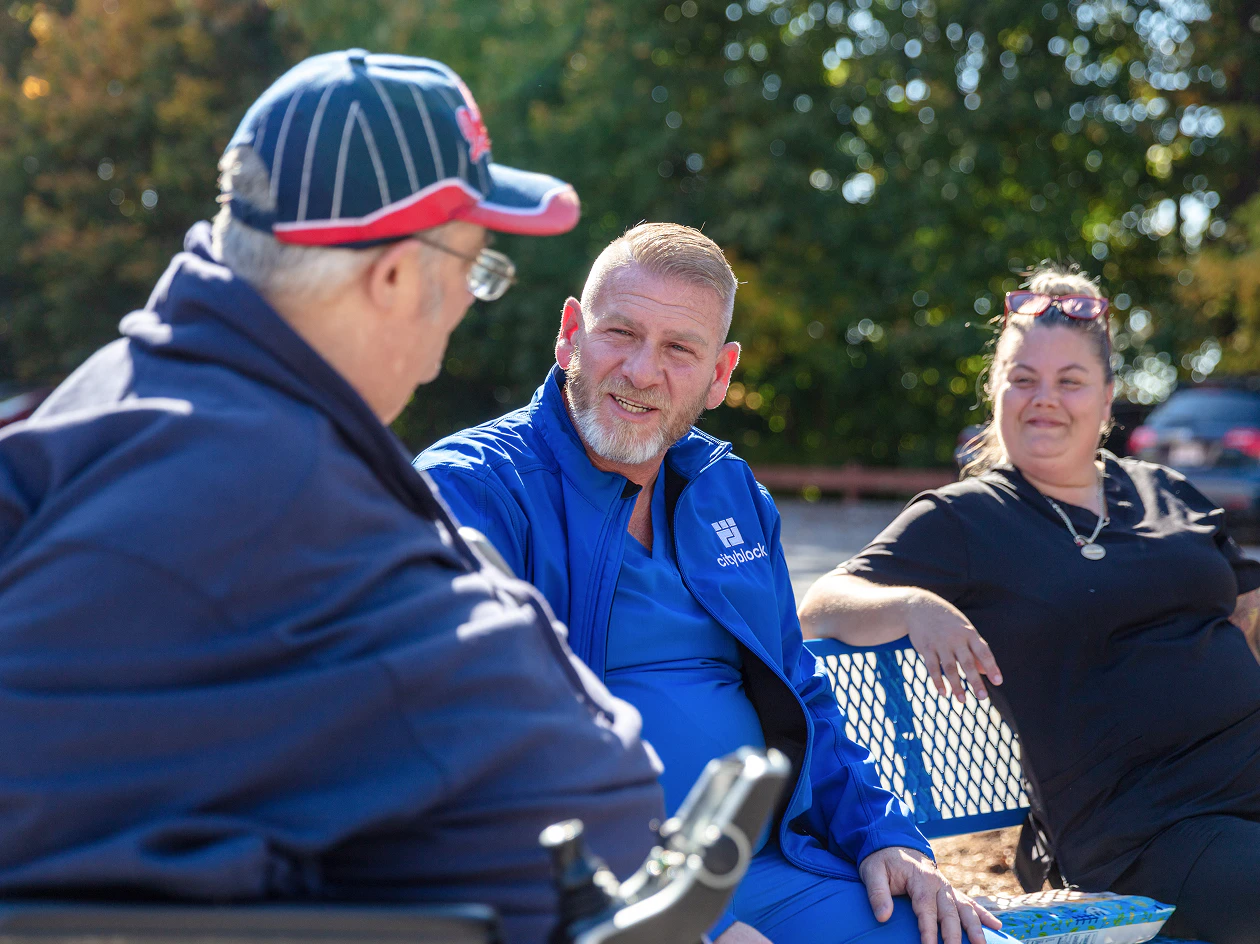 Image of a clinician in a blue jacket conversing outdoors with an older man sitting in a wheelchair, while a healthcare worker smiles nearby.