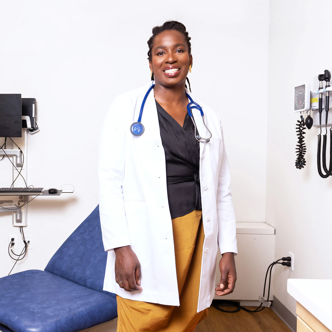 Image of Cityblock founder and CEO, Toyin Ajayi, wearing a white lab coat and blue stethoscope, standing in a medical examination room and smiling.