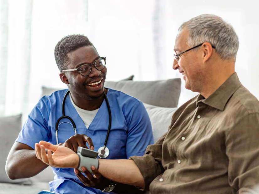 Image of a male healthcare worker in blue scrubs taking the blood pressure of an older man sitting on a sofa.