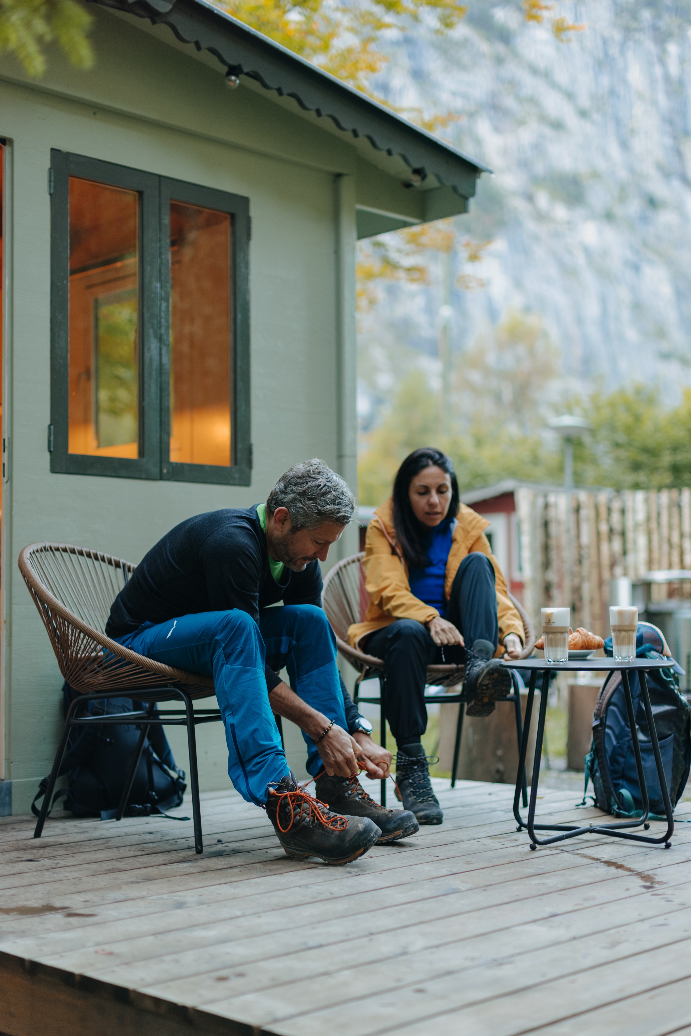Mann und Frau sitzen auf der Terrasse eines Fuchsbaus, eine Mietunterkunft auf Camping Jungfrau, und binden sich die Schuhe, um sich auf eine Wanderung vorzubereiten. 2 Kaffees stehen auf dem kleinen Tisch