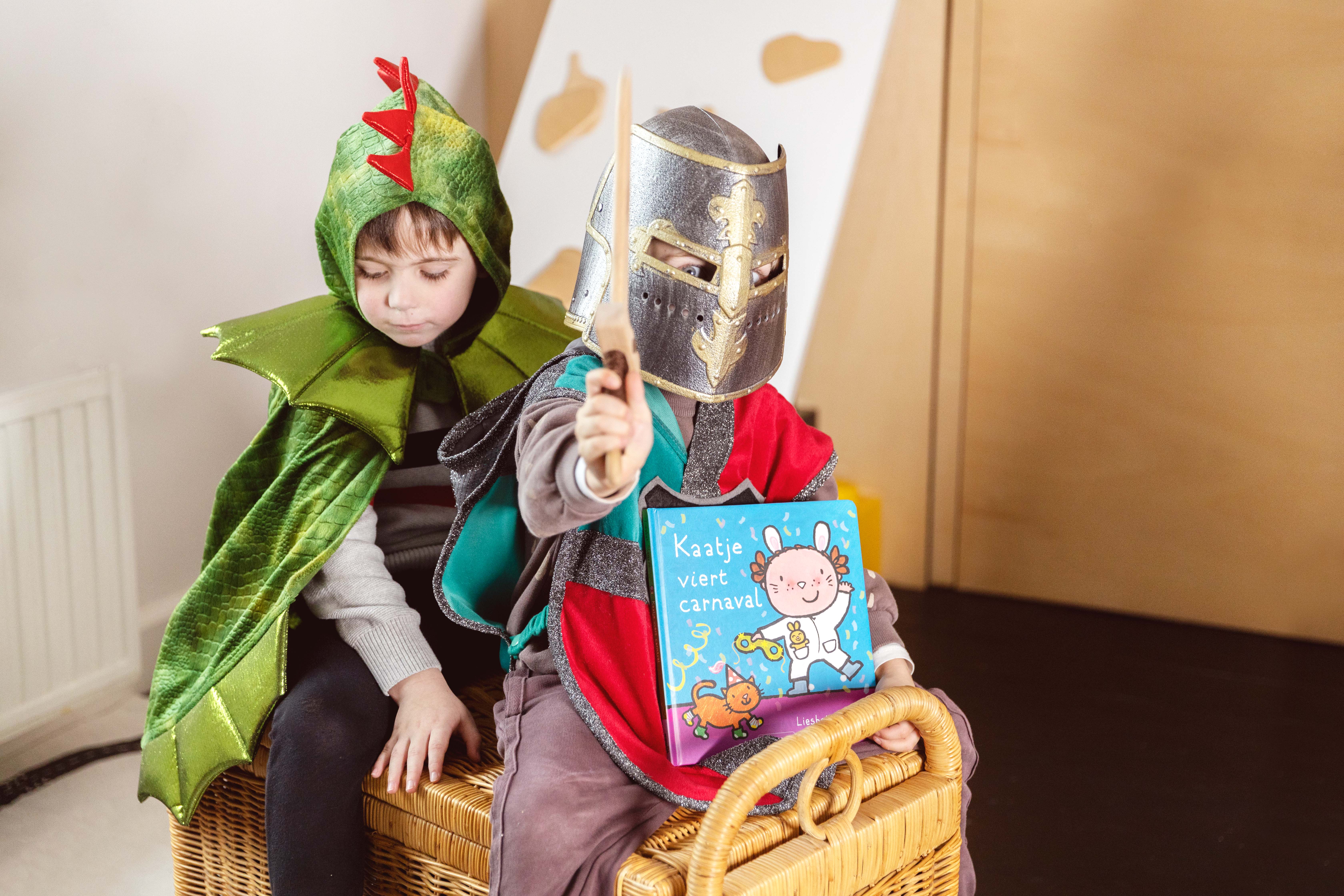 Foto van twee kleuters op een verkleed als draak en ridder op een verkleedkoffer met het carnavalsboek Kaatje viert carnaval van Liesbet Slegers.
