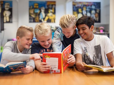 4 jongens in een klas die plezier beleven tijdens het lezen. Ze kijken samen in een leesboek van David Walliams en lachen.