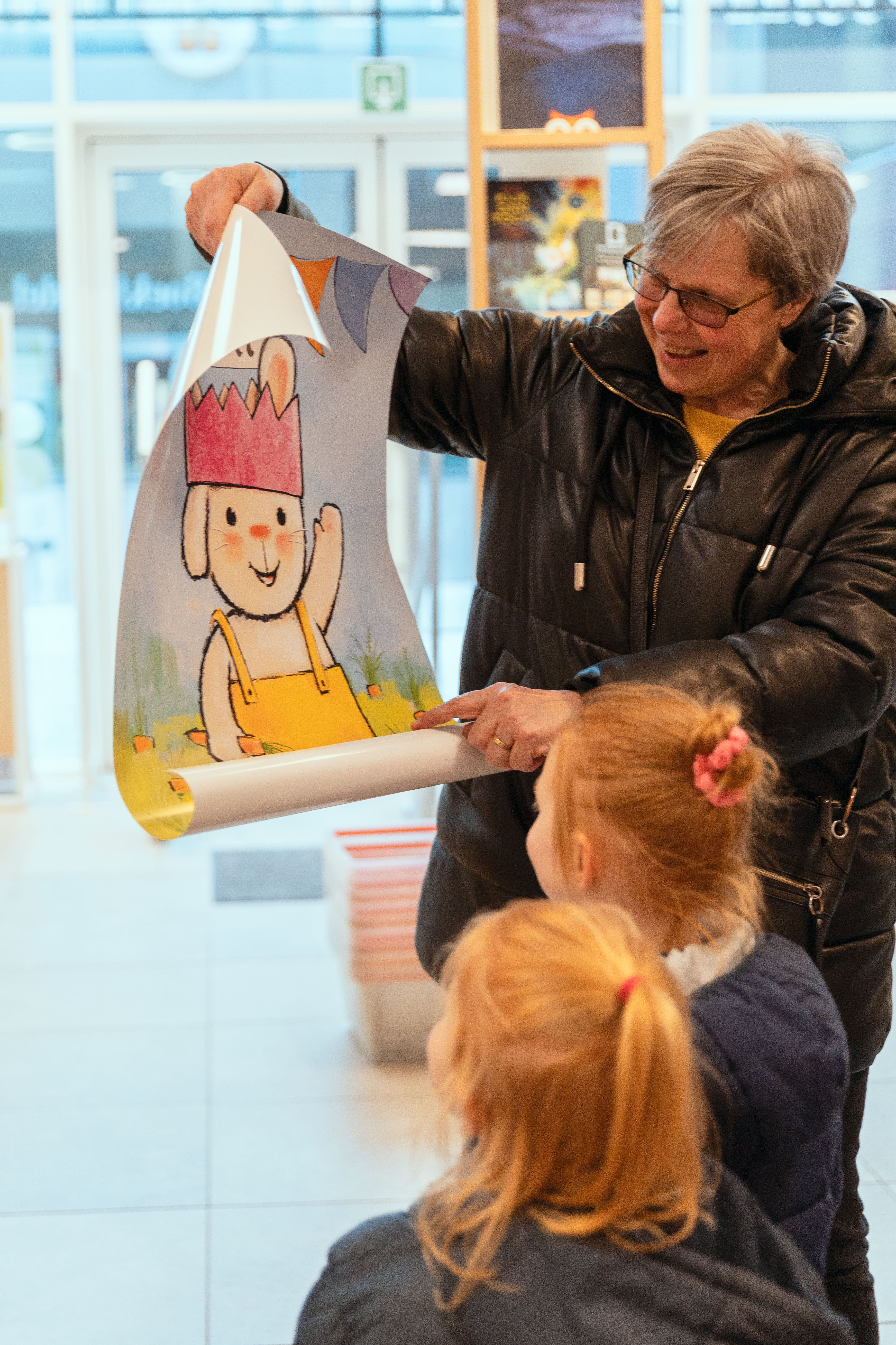 Een foto van een mevrouw die een poster van Rikki toont aan twee kindjes. 