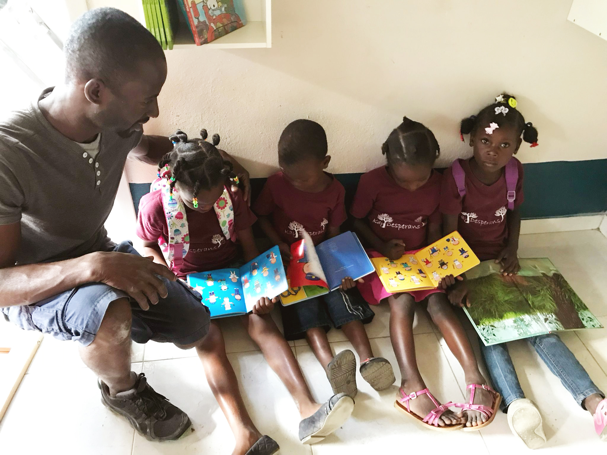 Een man zit op de grond naast vier jonge kinderen die boeken lezen. De kinderen dragen paarse schooluniformen en hebben kleurrijke rugzakken. Ze zitten op een betegelde vloer in een lichte ruimte met boekenplanken.