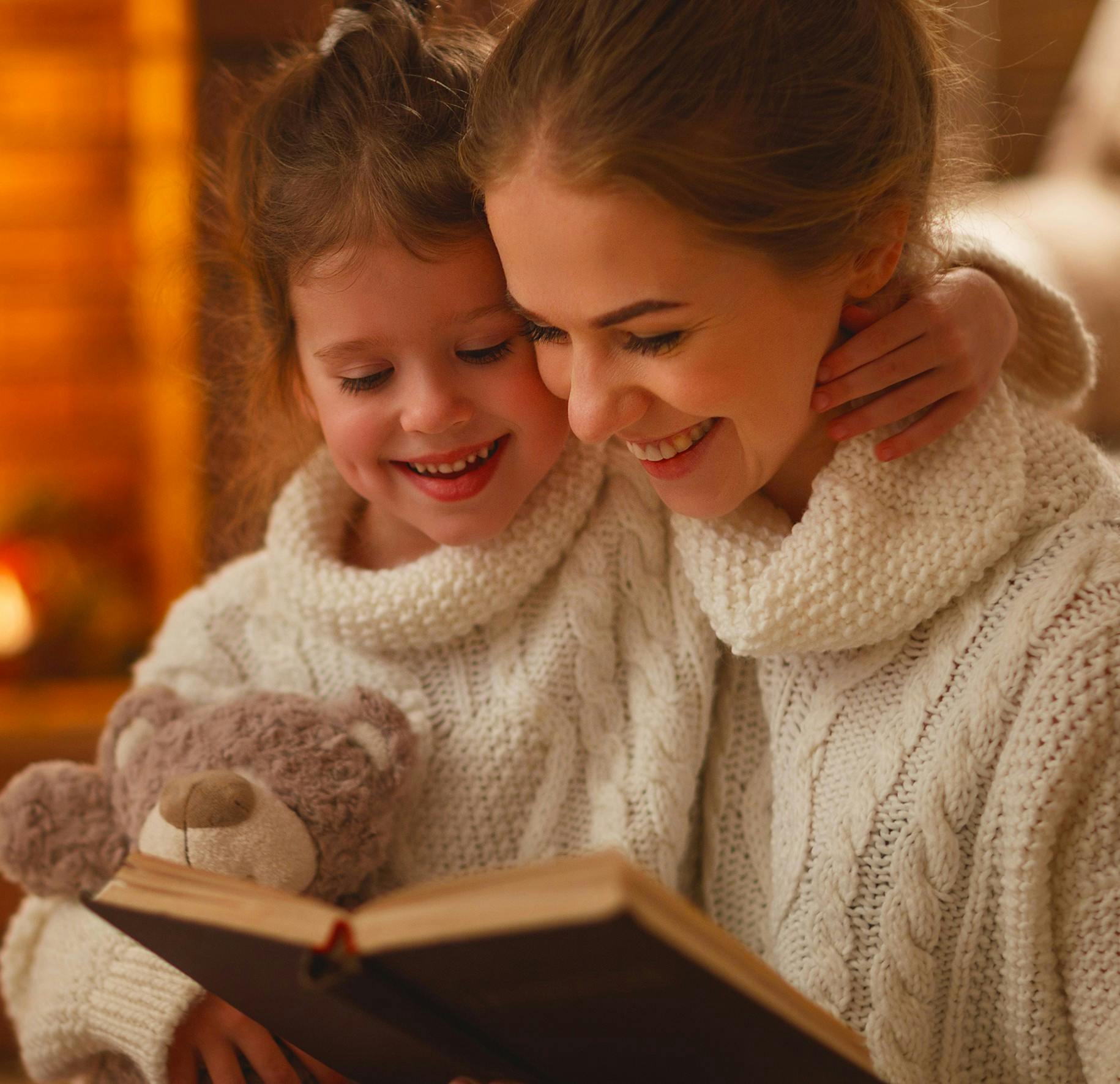 Foto van een moeder en haar dochter met warme wintertrui die allebei lachend in een boek kijken en samen genieten van een verhaal