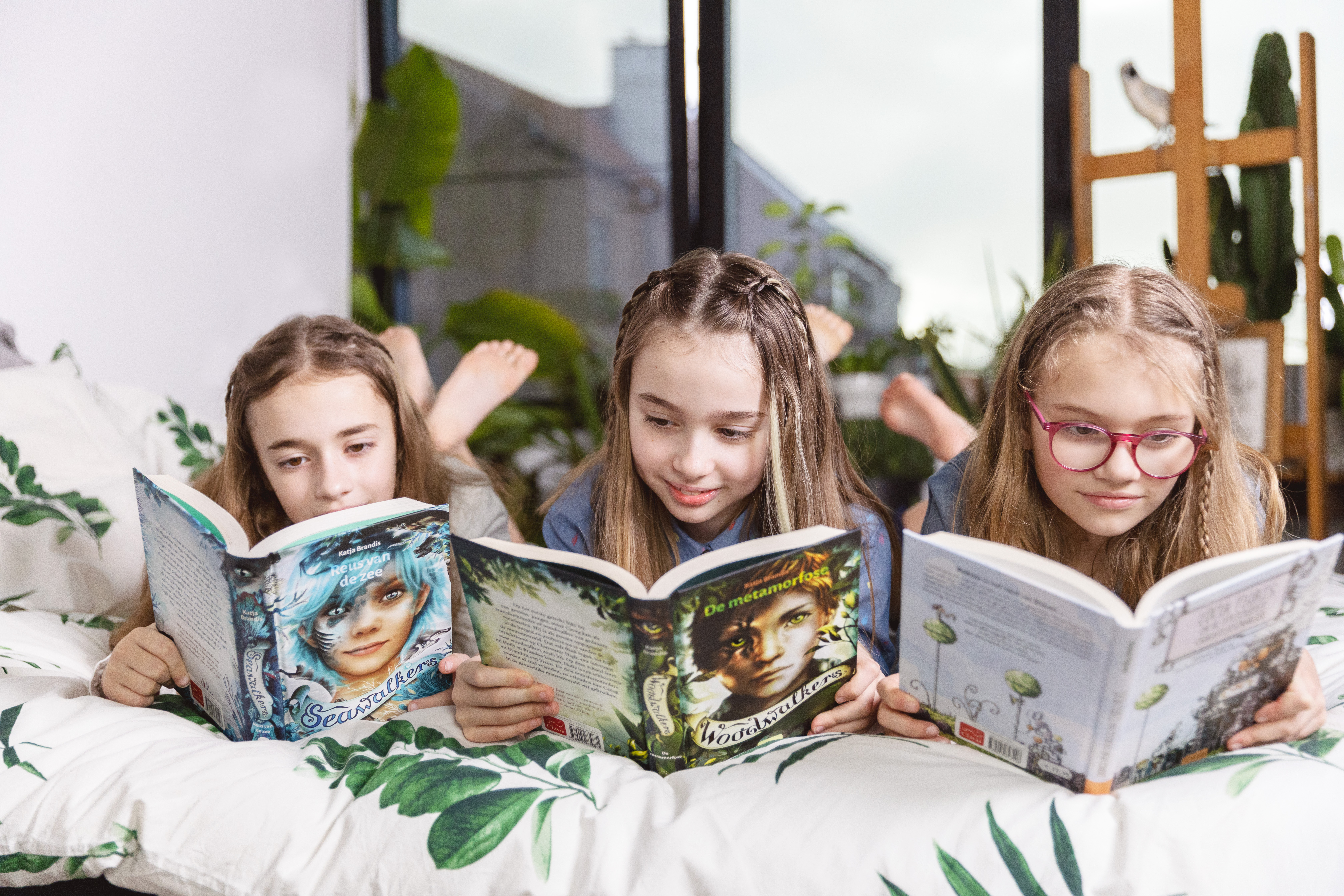 Drie meisjes liggen op hun buik op bed en ze lezen elk een boek.