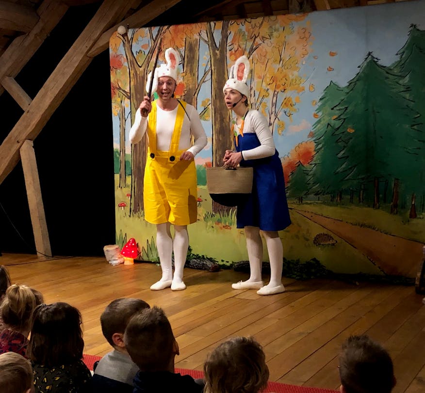 Een foto van de musical 'Ik ben Rikki'. We zien twee acteurs die Rikki en Anni spelen. Ze staan voor het decor staan. Het decor is een doek waarop een bos te zien is. Er zitten enkele kindjes op de mat naar de musical de kijken. 