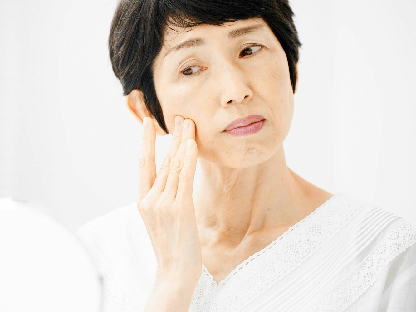 An older woman touches her face while looking at her reflection in a hand mirror