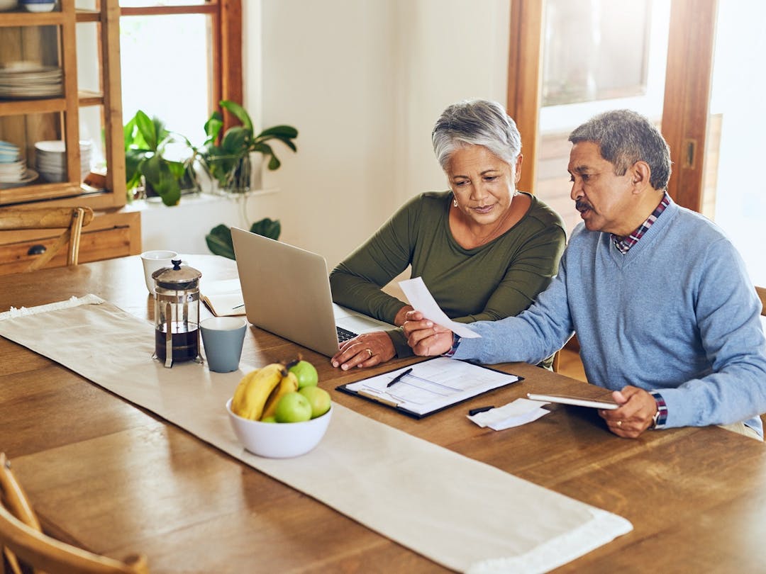 An older Hispanic husband and wife sit at the table in front of a laptop and do their taxes