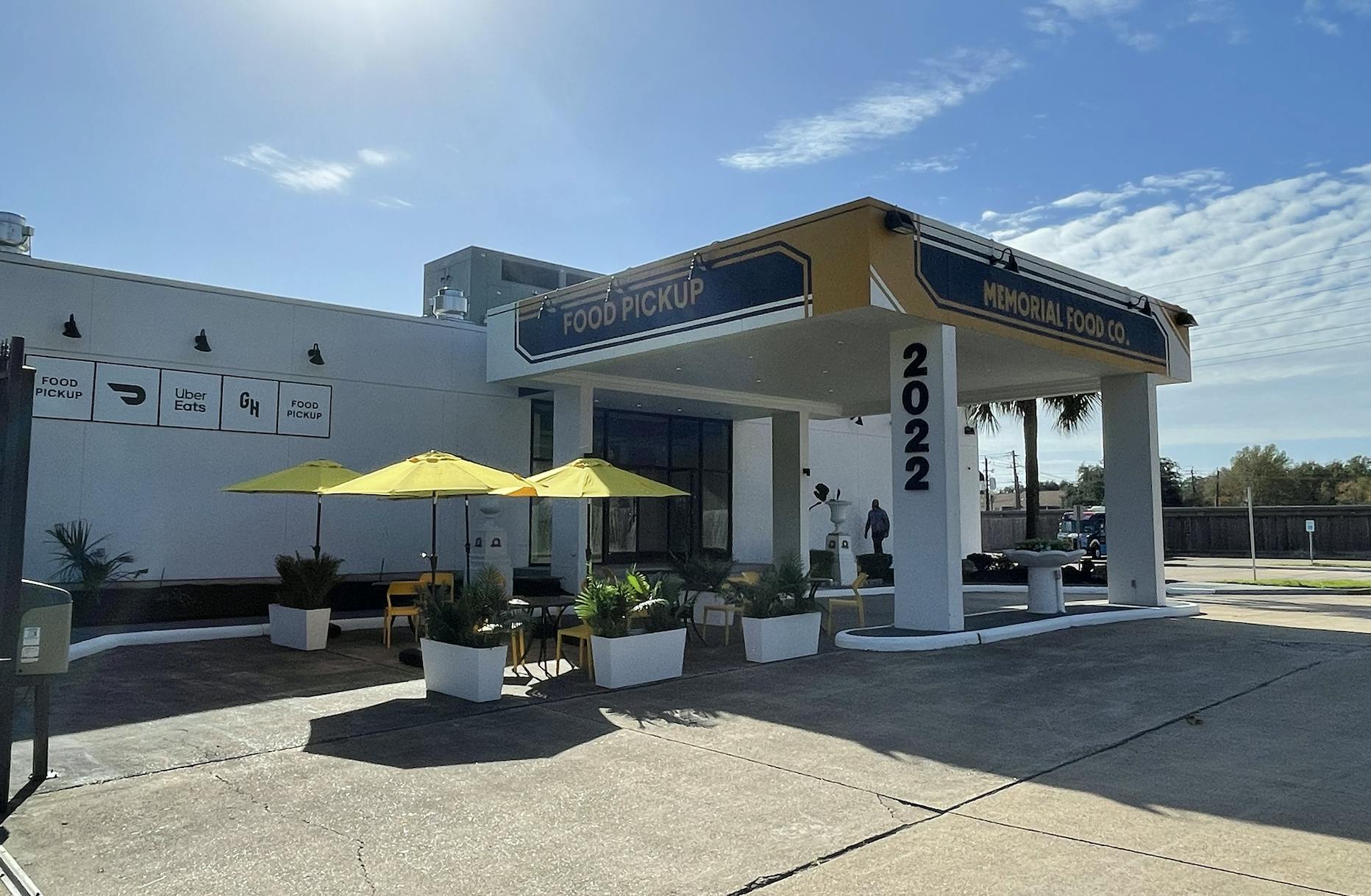 White facility with signs of delivery services on the wall, and two bigger signs with "Food Pickup" and "Memorial Food Co." written, tables with sun-shades, chairs and decorative plants, on the back there is a little area of vegetation and there aren't many clouds in the sky and it's sunny