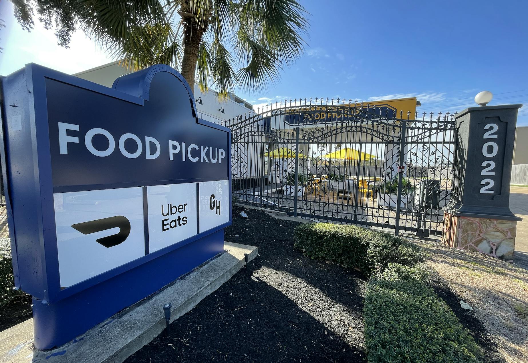 Blue sign with "Food PIckup" written in white on a little garden on the entrance of a facility, that is behind a grey gate