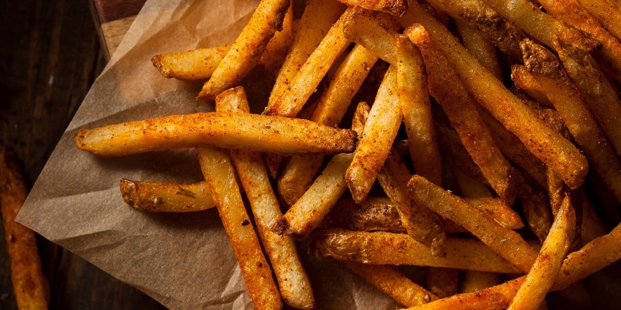 A fries portion served on a wooden board lined with some paper, on the back, behind the board, is a darker wood