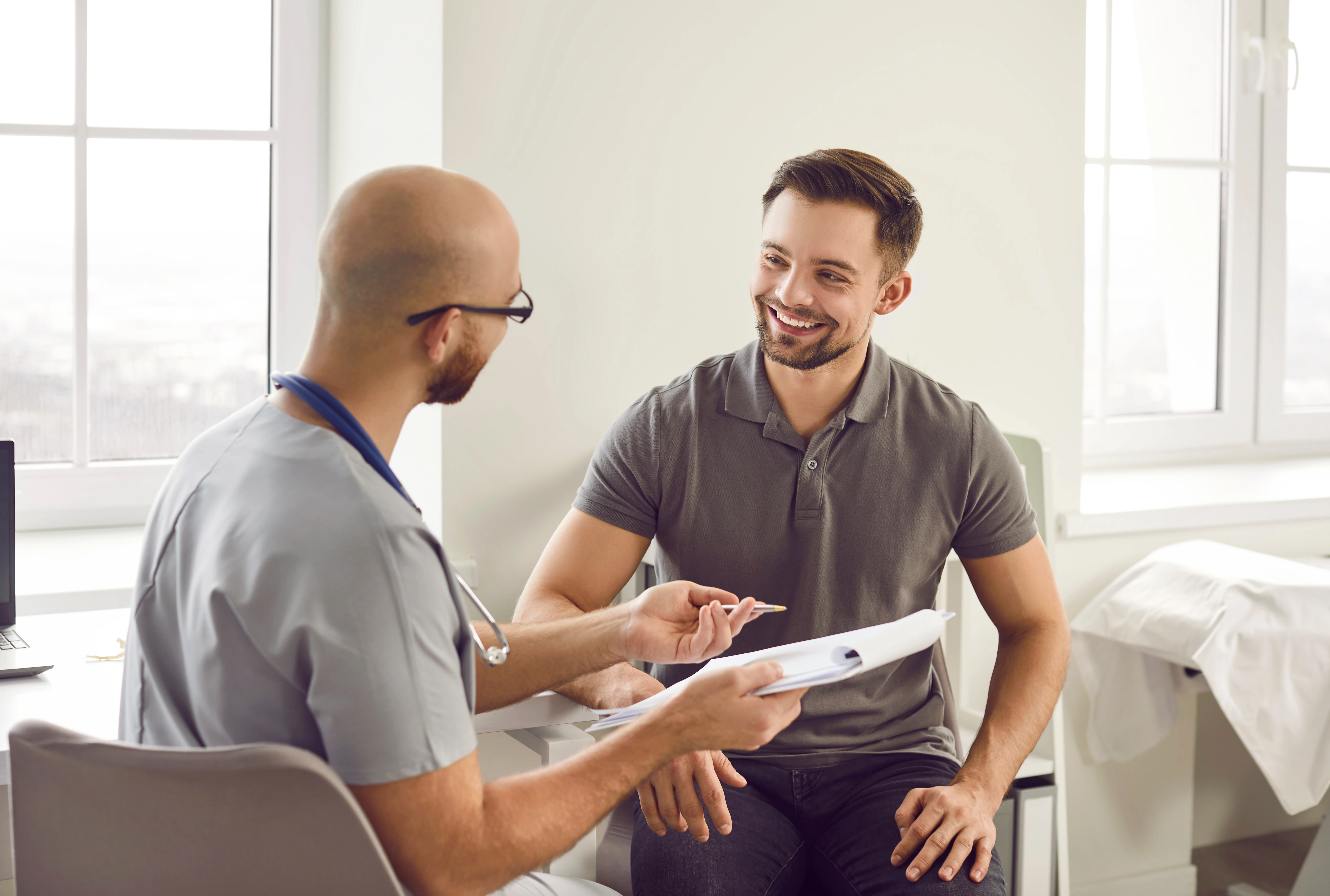 A male patient is looking happily at his male doctor holding a clipboard. 