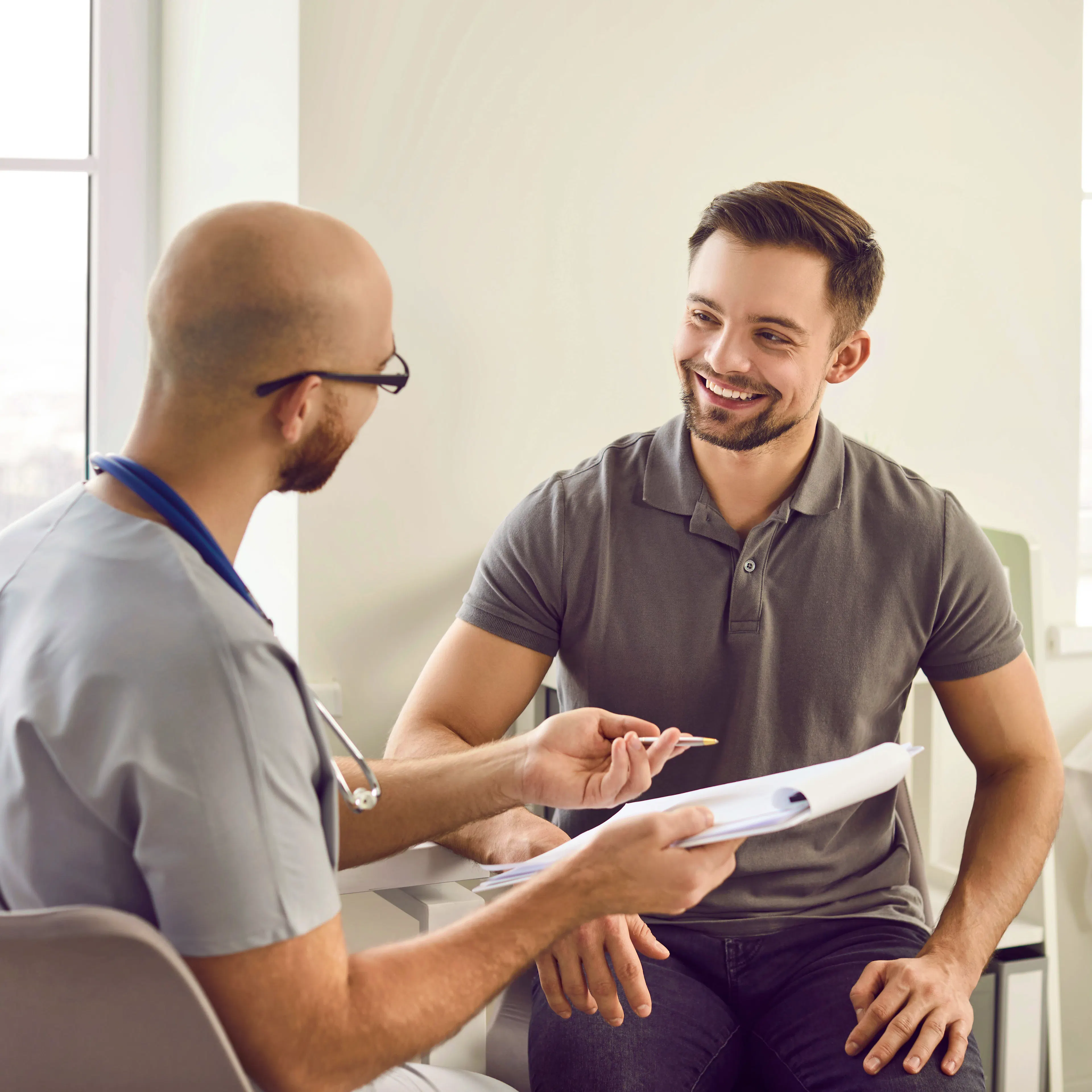 A male patient is looking happily at his male doctor holding a clipboard.