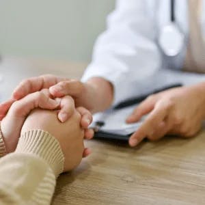 A healthcare provider in a white coat and a stethoscope hanging around her right shoulder embraces the hand of a patient.