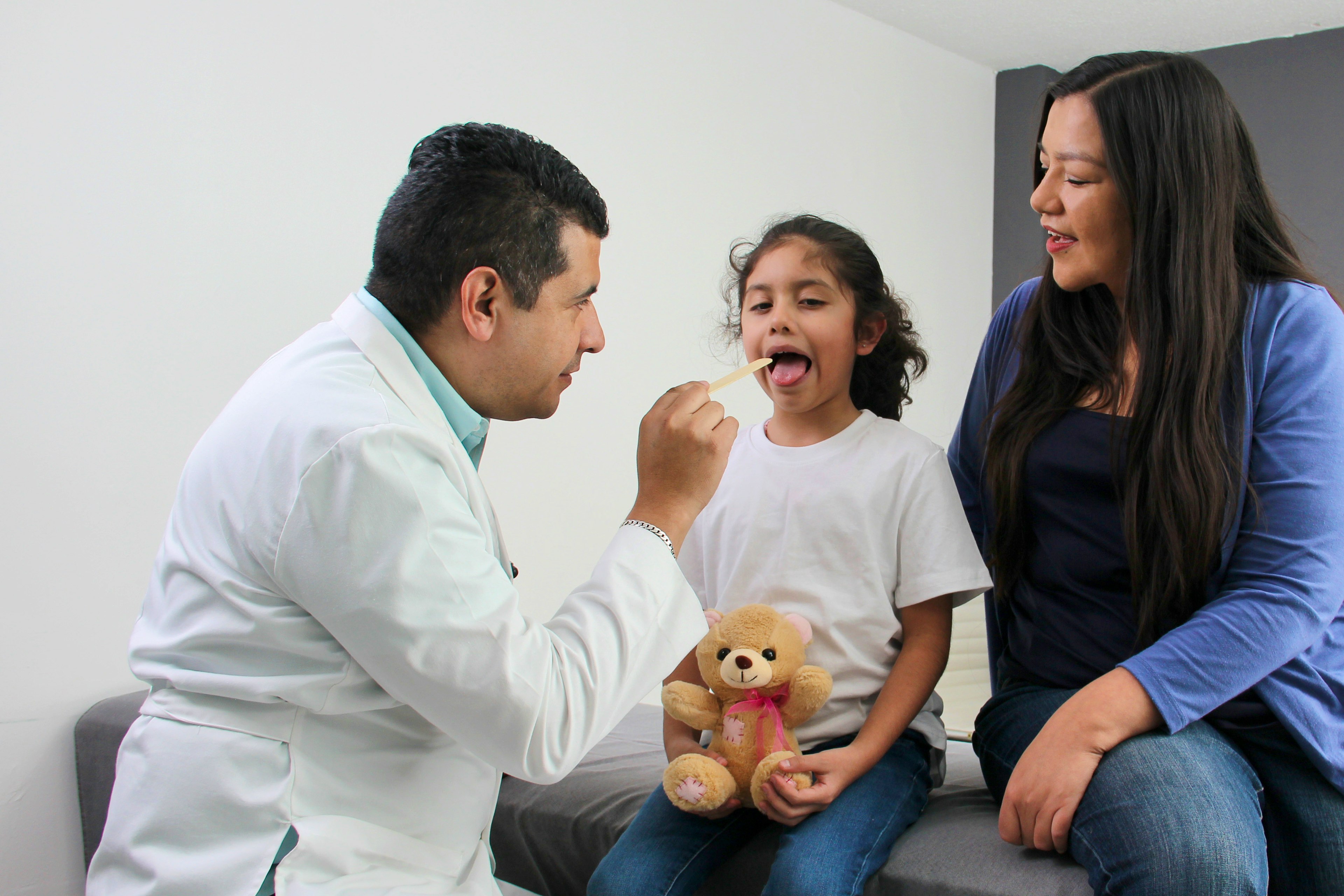 A physician examines child's throat with a tongue depressor as her mother observes.