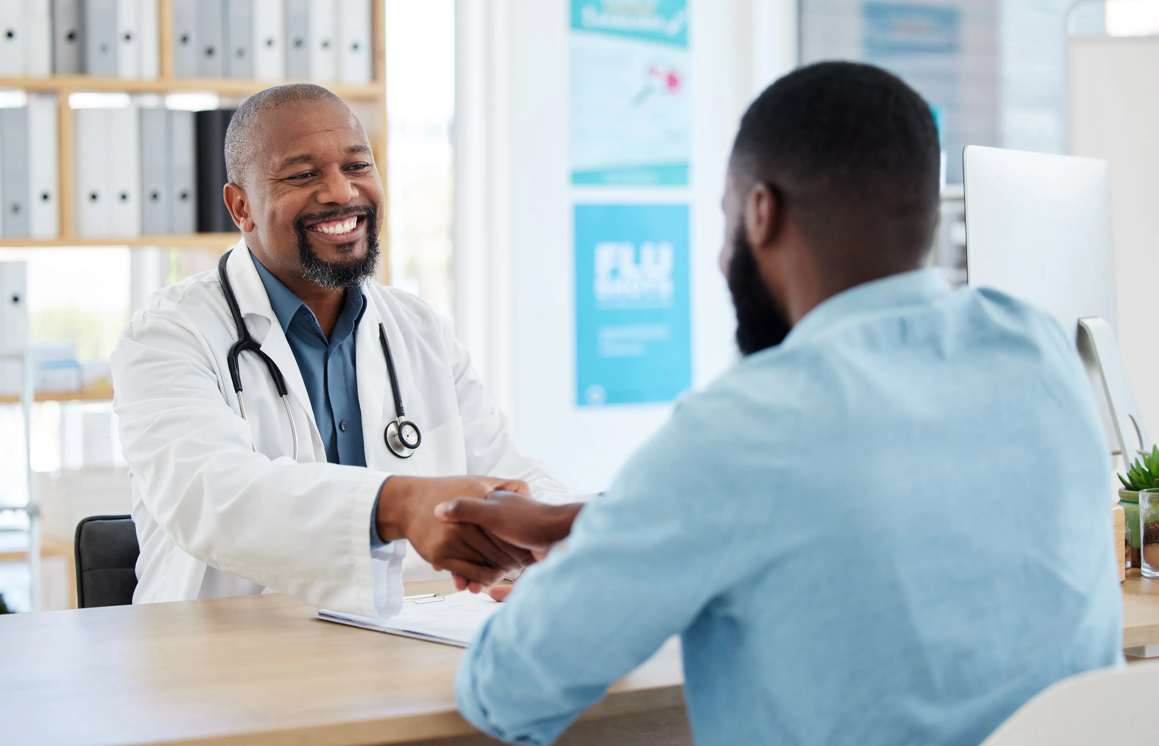A male doctor in his white coat is shaking a male patient's hand at his desk. 