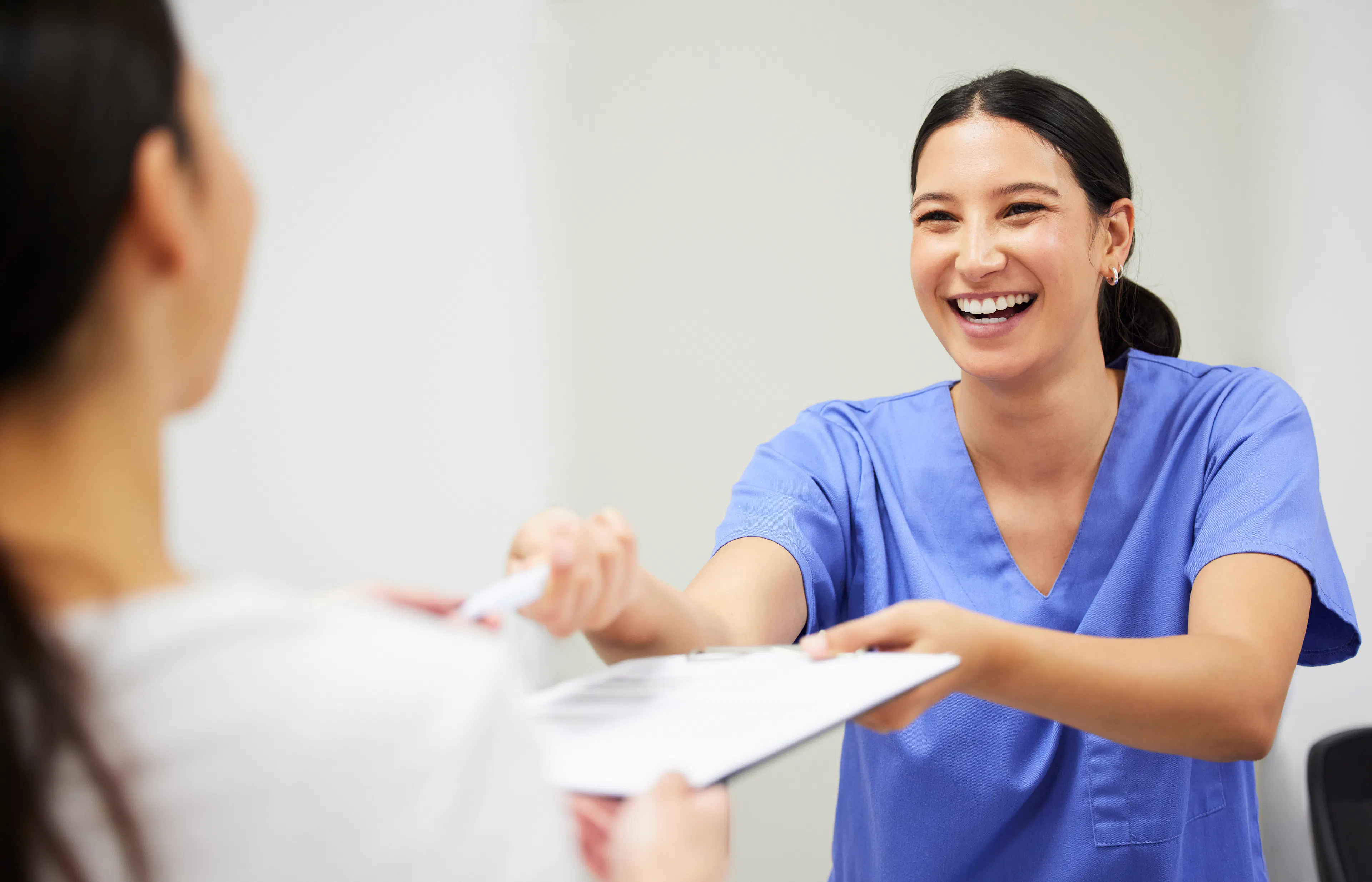 A medical worker in scrubs is smiling at a patient, handing her paperwork to fill out.