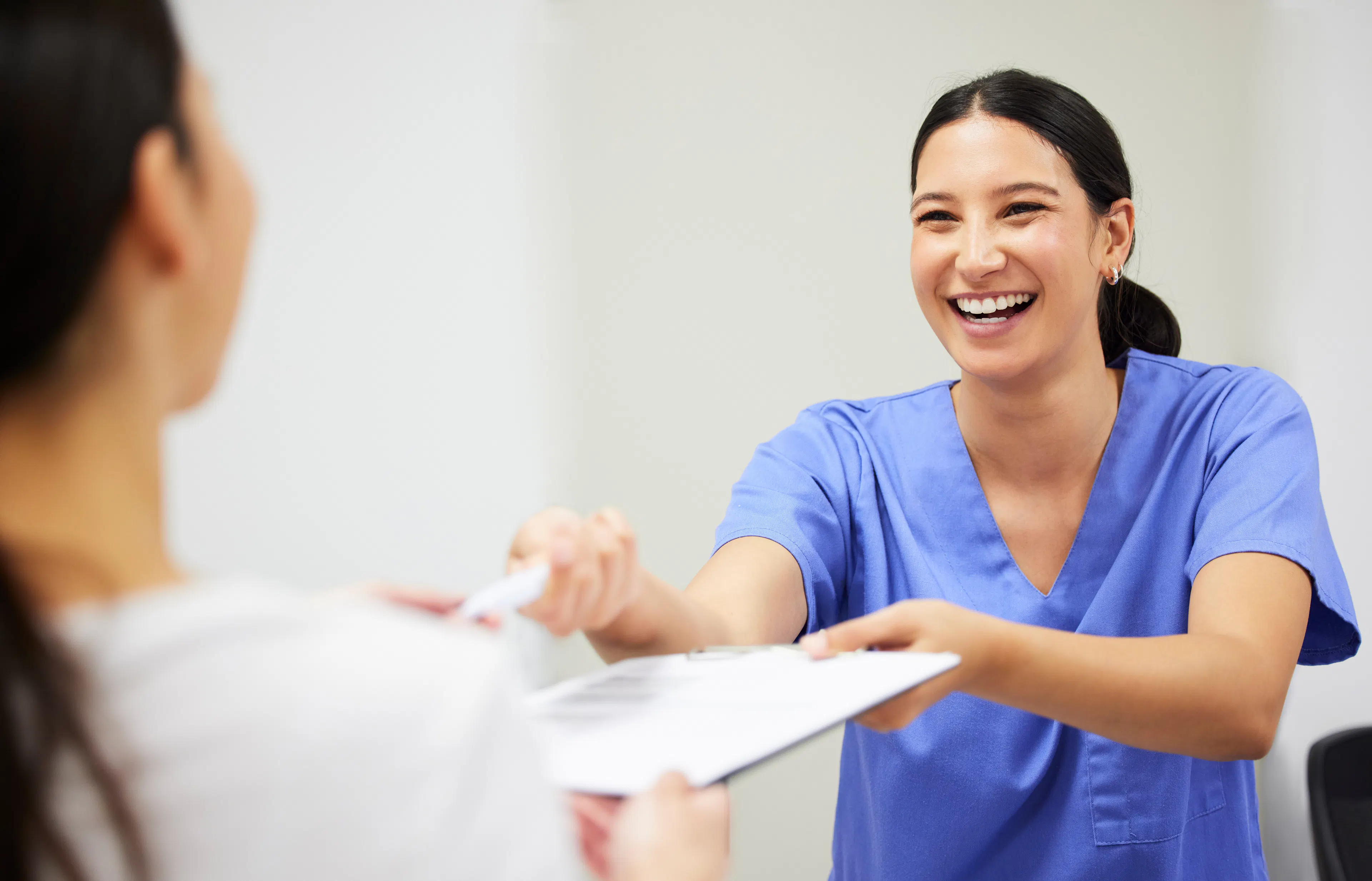 A medical worker in scrubs is smiling at a patient, handing her paperwork to fill out. 
