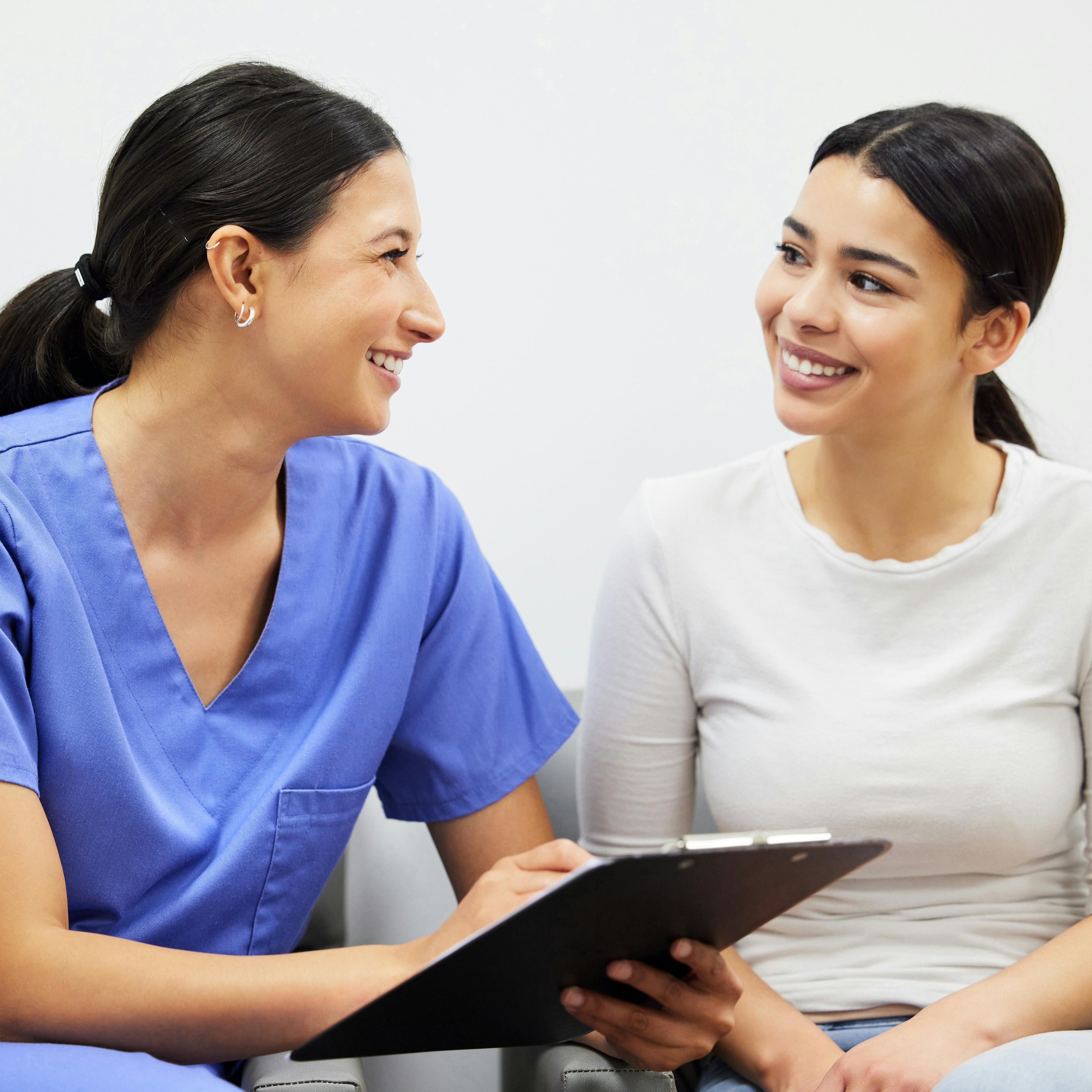 A medical worker in scrubs is sitting next to a patient, reviewing paperwork. Both women are smiling.  