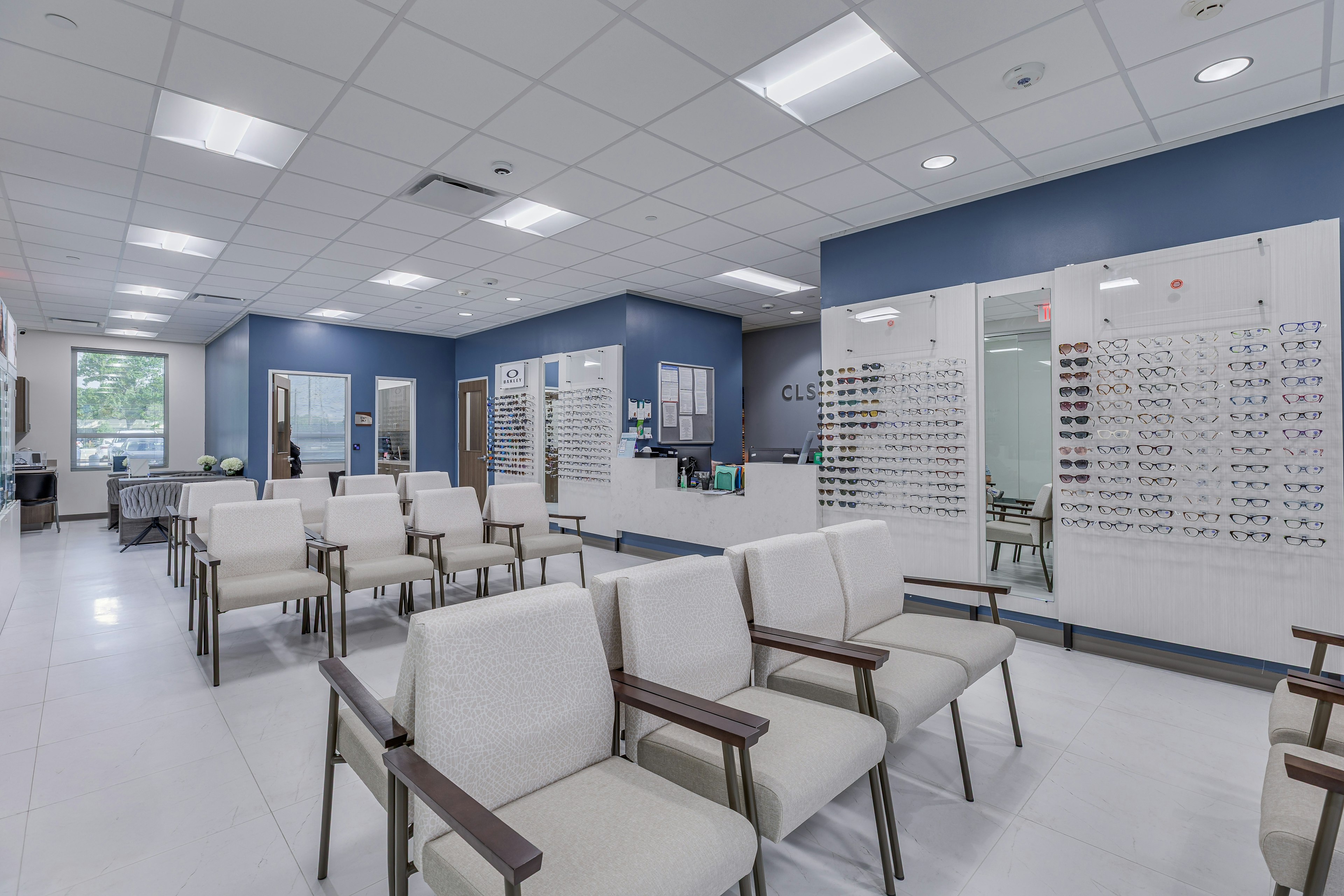 Modern optical waiting area with rows of upholstered chairs, a reception desk labeled "CLS Health," and a large wall display featuring a variety of eyeglass frames. The space is clean, bright, and welcoming, with blue accent walls and ample natural light.