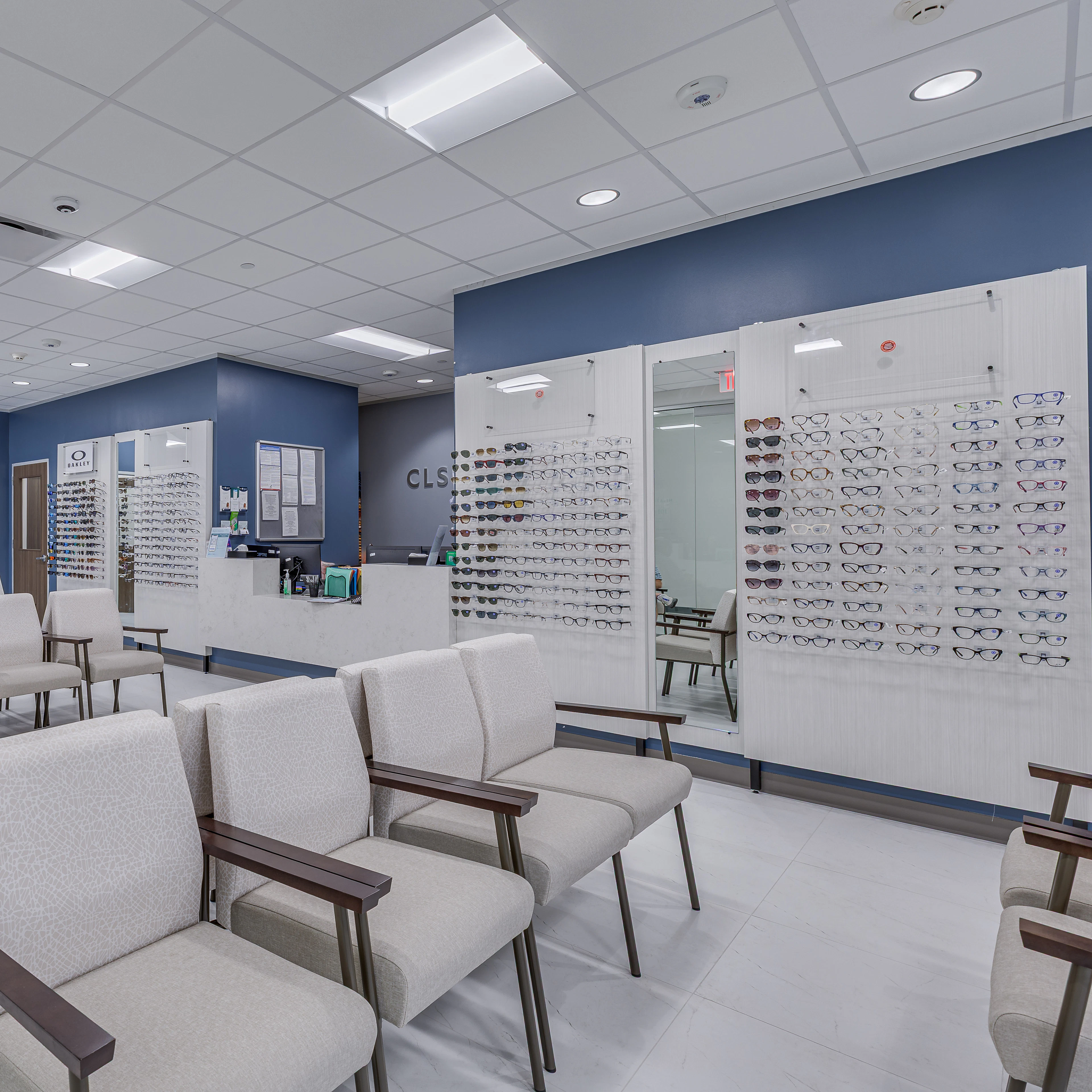 Modern optical waiting area with rows of upholstered chairs, a reception desk labeled "CLS Health," and a large wall display featuring a variety of eyeglass frames. The space is clean, bright, and welcoming, with blue accent walls and ample natural light.