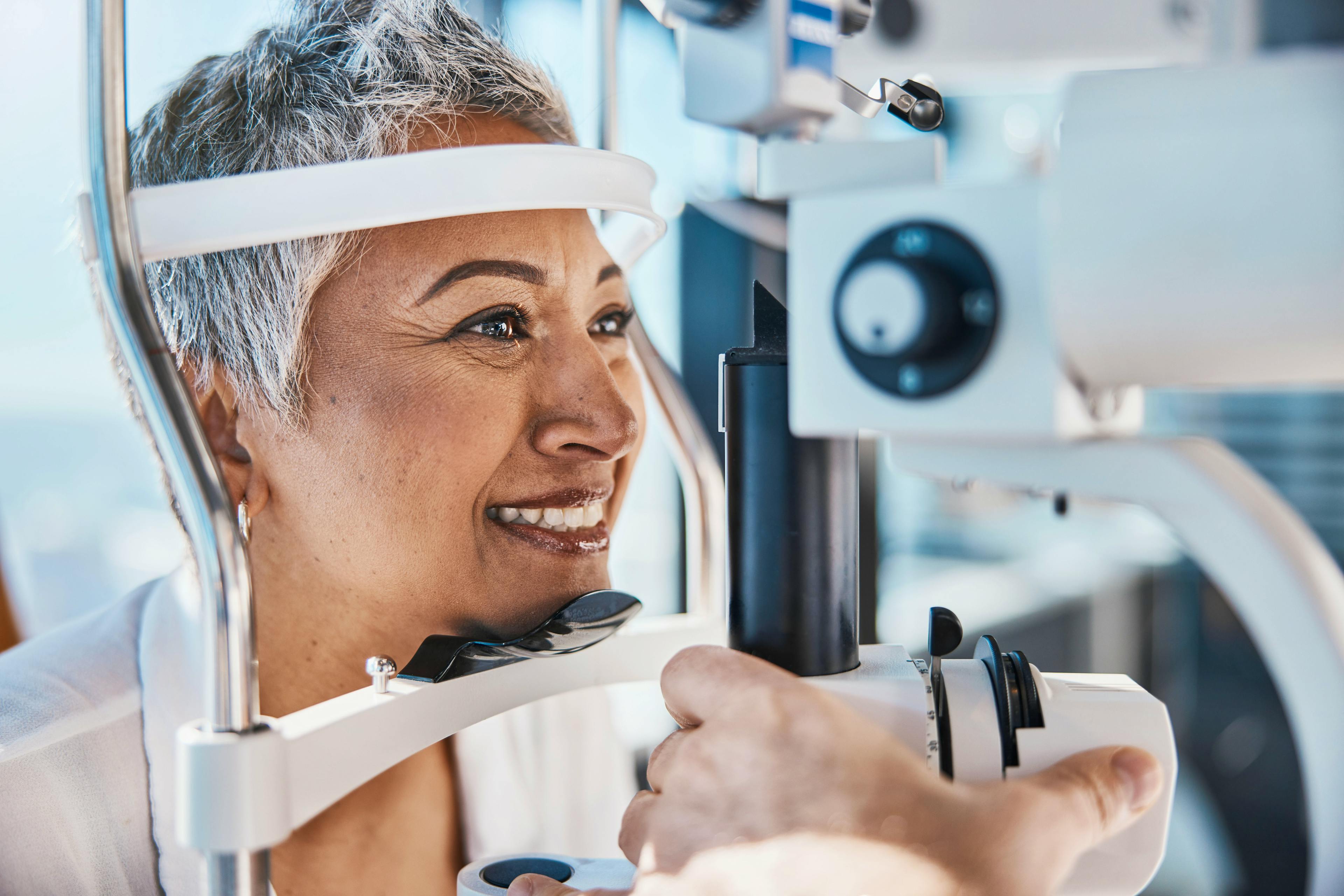 Mature woman with short gray hair smiles during an eye examination, positioned at a slit lamp in a well-lit clinic, as a healthcare provider adjusts the instrument.