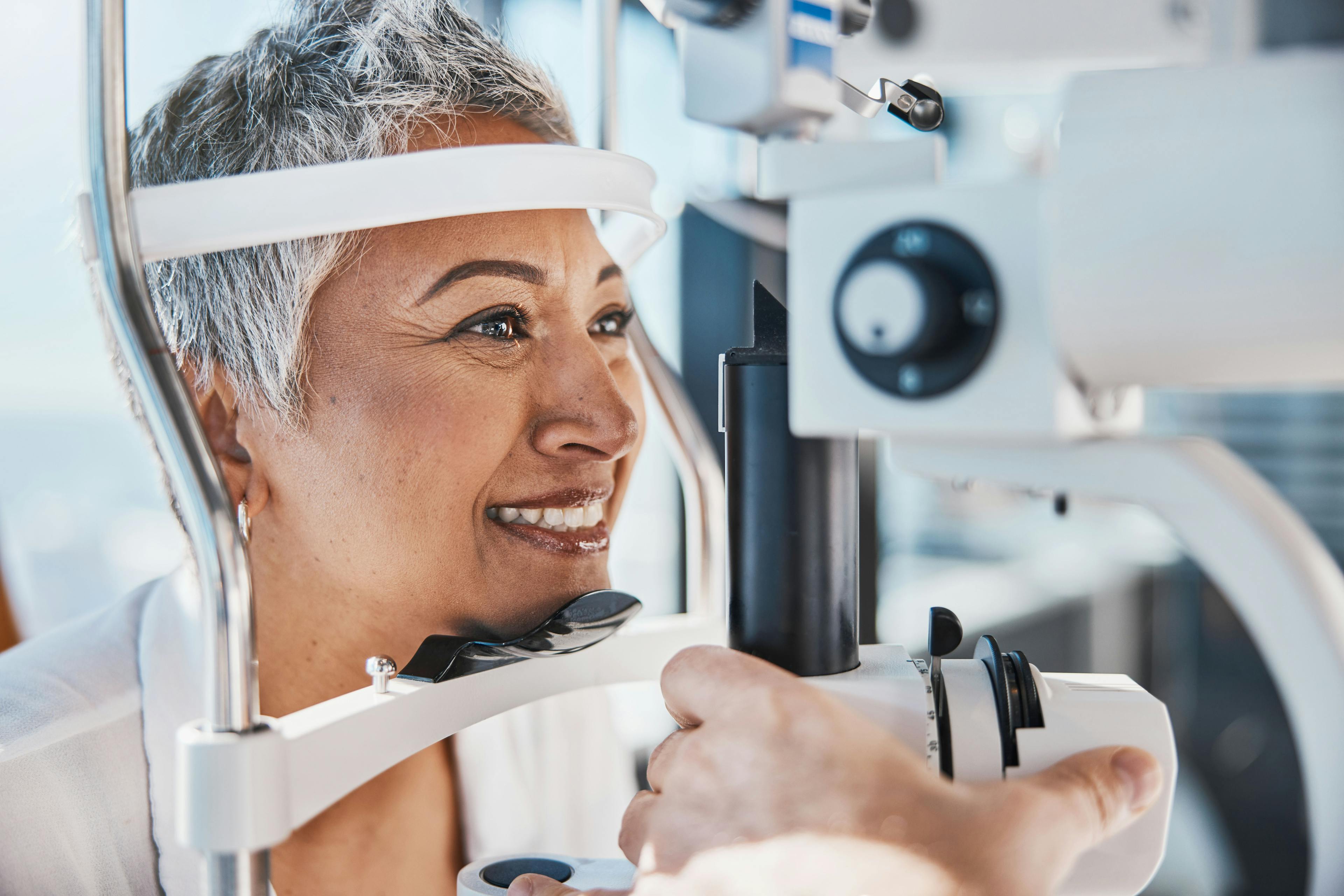 Mature woman with short gray hair smiles during an eye examination, positioned at a slit lamp in a well-lit clinic, as a healthcare provider adjusts the instrument.