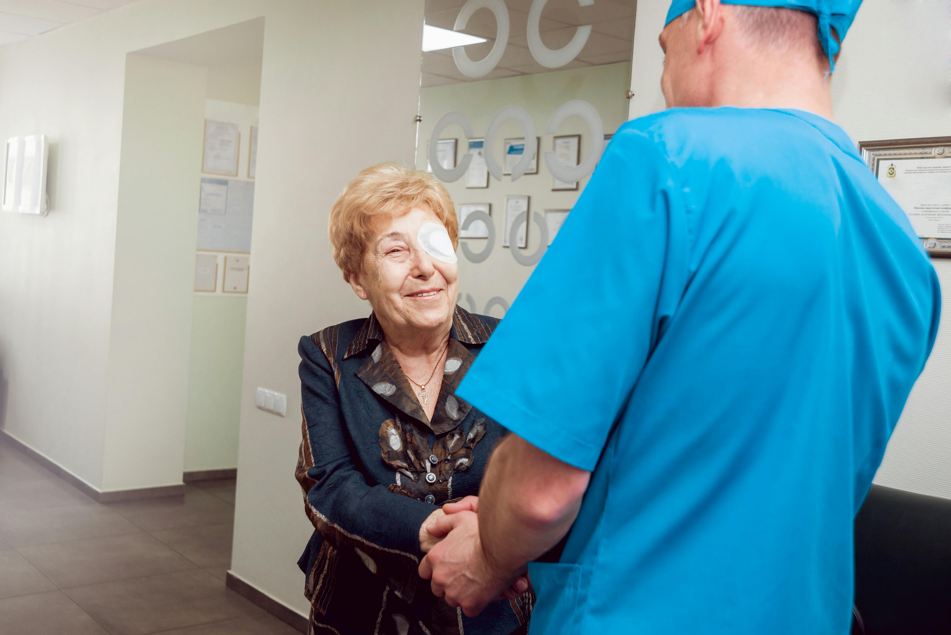 Smiling older woman with an eye patch shakes hands with a medical professional in scrubs, likely after cataract surgery, standing in a bright, modern clinic hallway