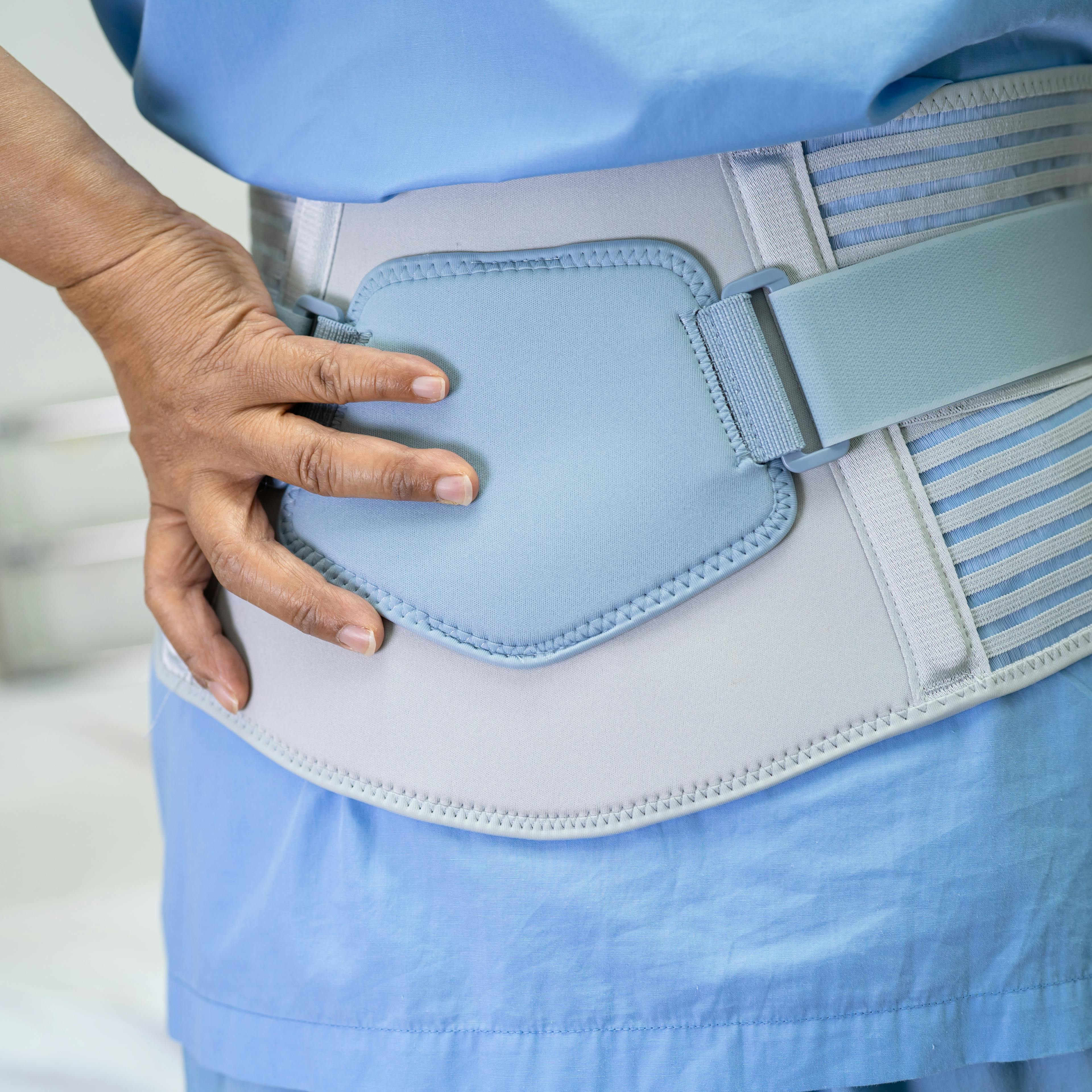 A person wearing a medical back brace places their hand on their lower back, indicating pain or discomfort, in a clinical setting.