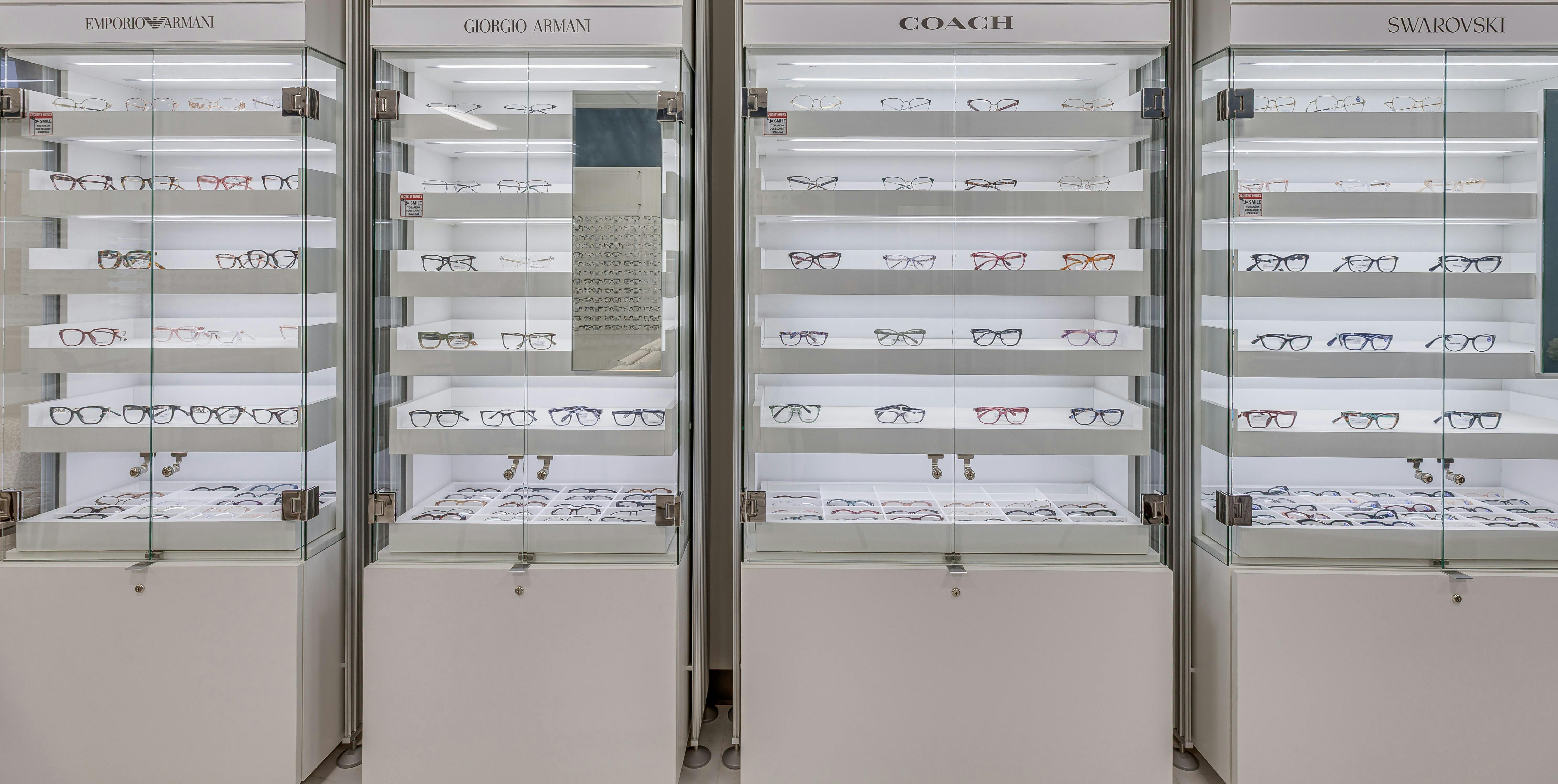 Glass display cases showcasing designer eyeglass frames from Emporio Armani, Giorgio Armani, Coach, and Swarovski, each labeled with brand signage and promotional images above the shelves. The cases are well-lit, with multiple rows of glasses neatly arranged on transparent shelves.