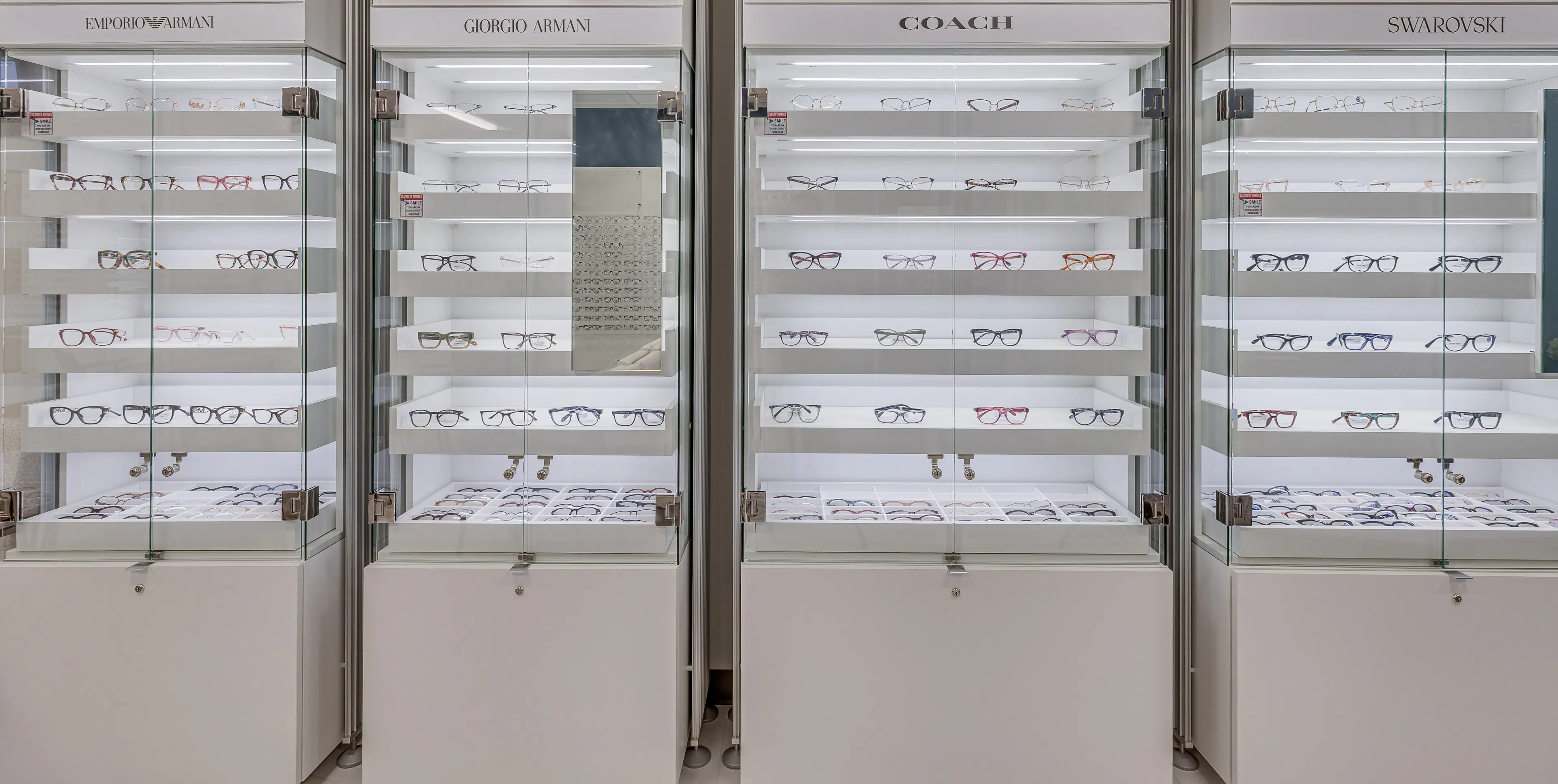 Glass display cases showcasing designer eyeglass frames from Emporio Armani, Giorgio Armani, Coach, and Swarovski, each labeled with brand signage and promotional images above the shelves. The cases are well-lit, with multiple rows of glasses neatly arranged on transparent shelves.