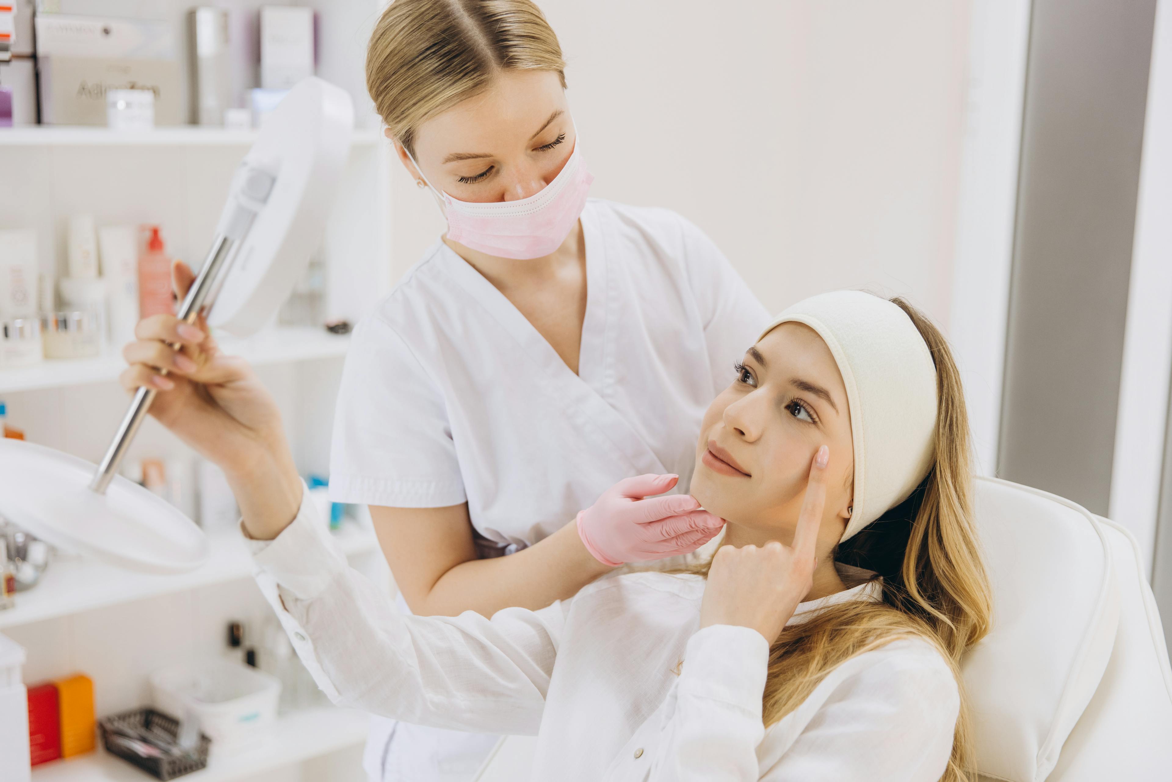 A cosmetics patient is observing her face with a mirror, with her provider helping.
